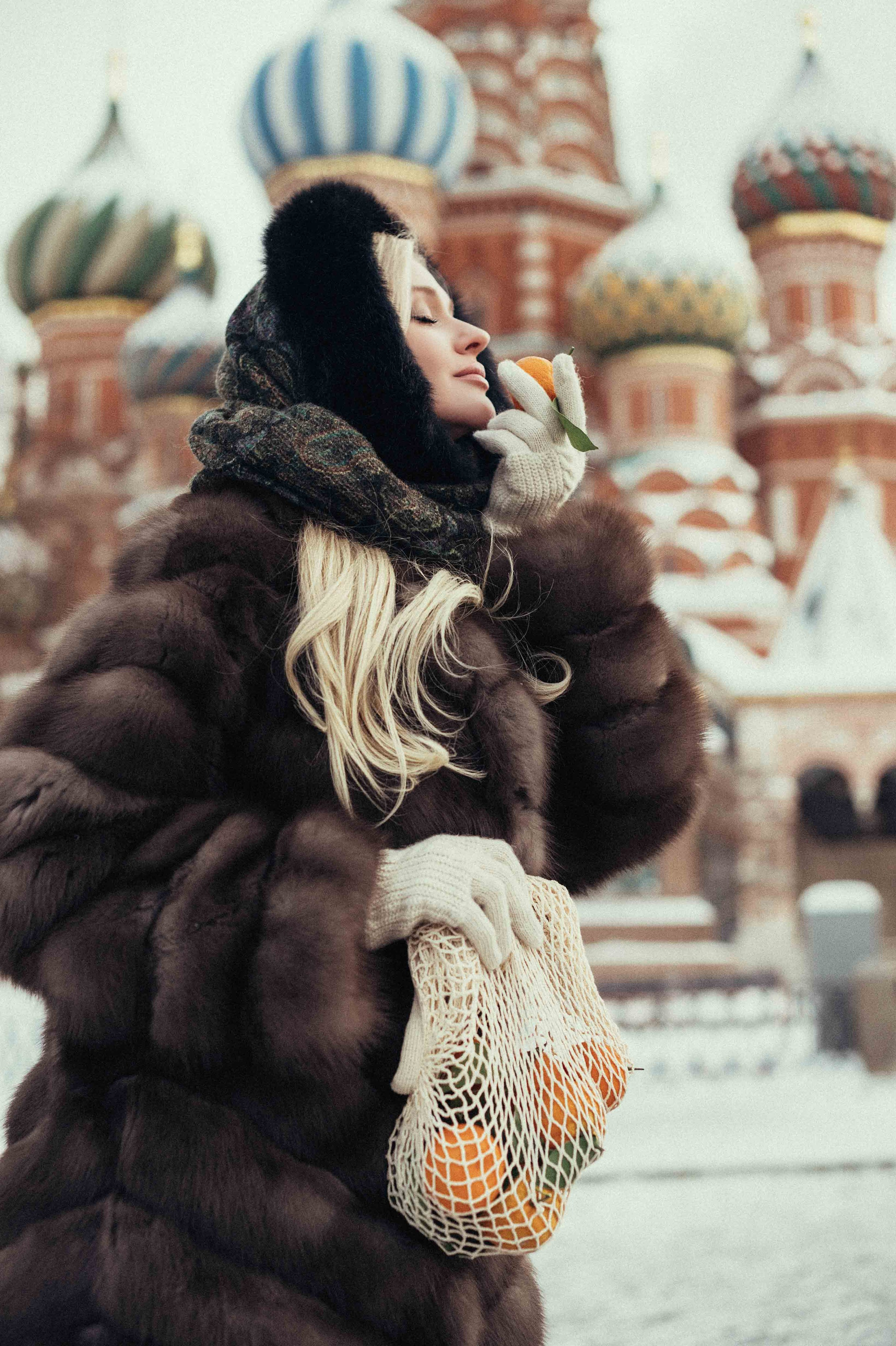 Девушка в стиле Russian girl  крупным планом на фоне куполов храма Василия Блаженного на Красной площади в Москве, стоит в профиль; в правой руке у нее авоська с мандаринами, а в левой один мандарин