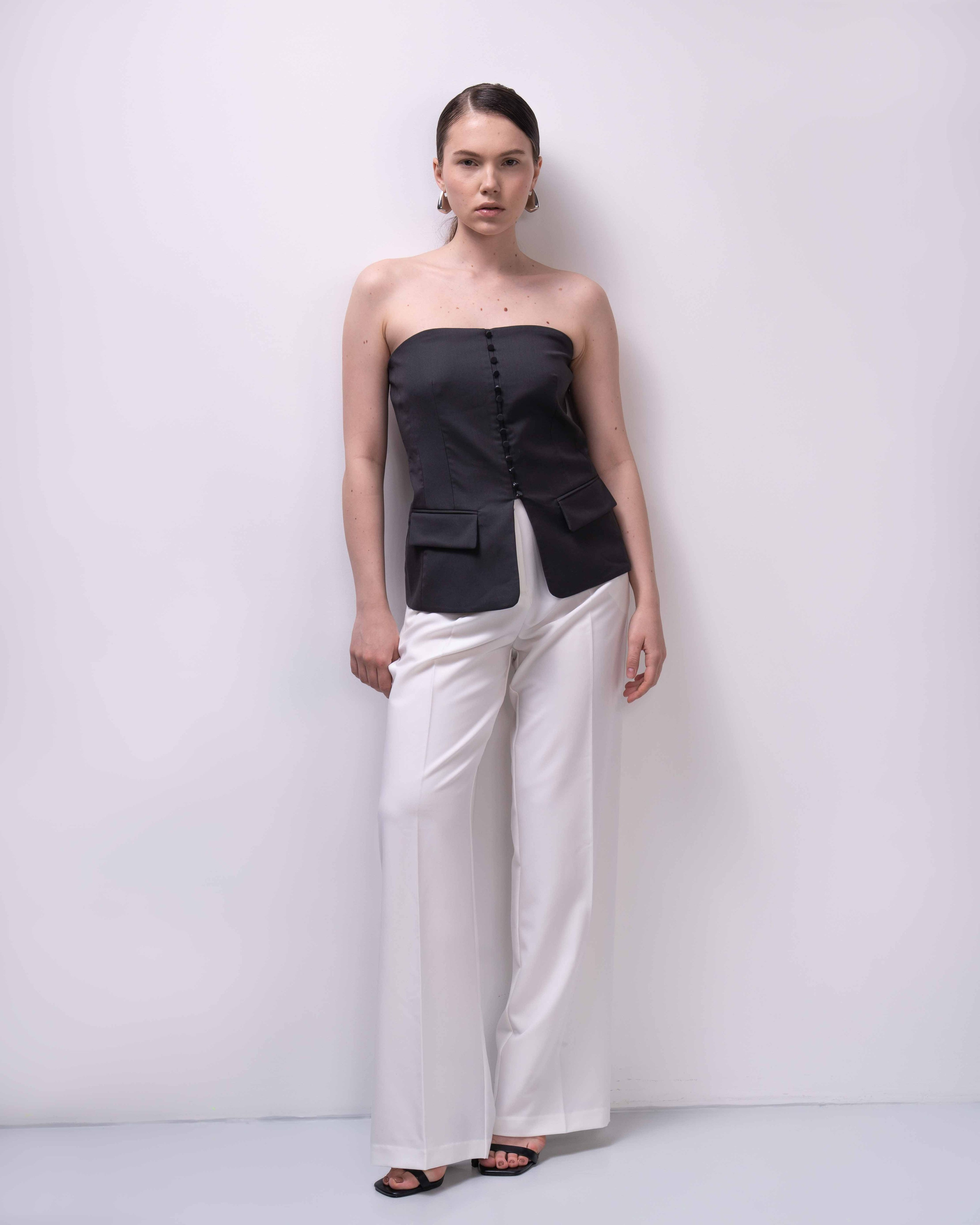 Фото товара для каталога  Ozon или Wildberries.  На фотографии изображена женщина, стоящая на светлом фоне. Она одета в черный топ без рукавов, который имеет свободный крой и застегивается на пуговицы спереди. Брюки у нее белые, прямого кроя, с высокой талией. На ногах у женщины черные сандалии с ремешками. Ее волосы собраны в аккуратный пучок, а на ушах видны серьги. Женщина смотрит прямо в камеру, ее поза расслабленная, руки опущены вдоль тела.