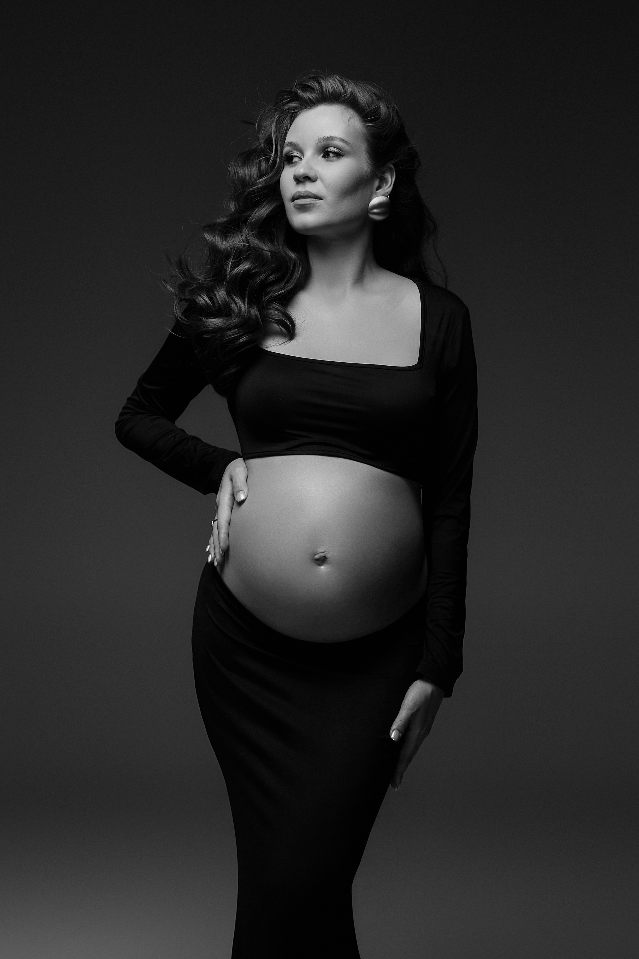 Беременная женщина в черном облегающем топе и юбке, стоит в студии и смотрит вниз на свой живот, нежно обнимая его руками. Это черно-белое изображение, подчеркивающее силуэт и свет. фотографсургут