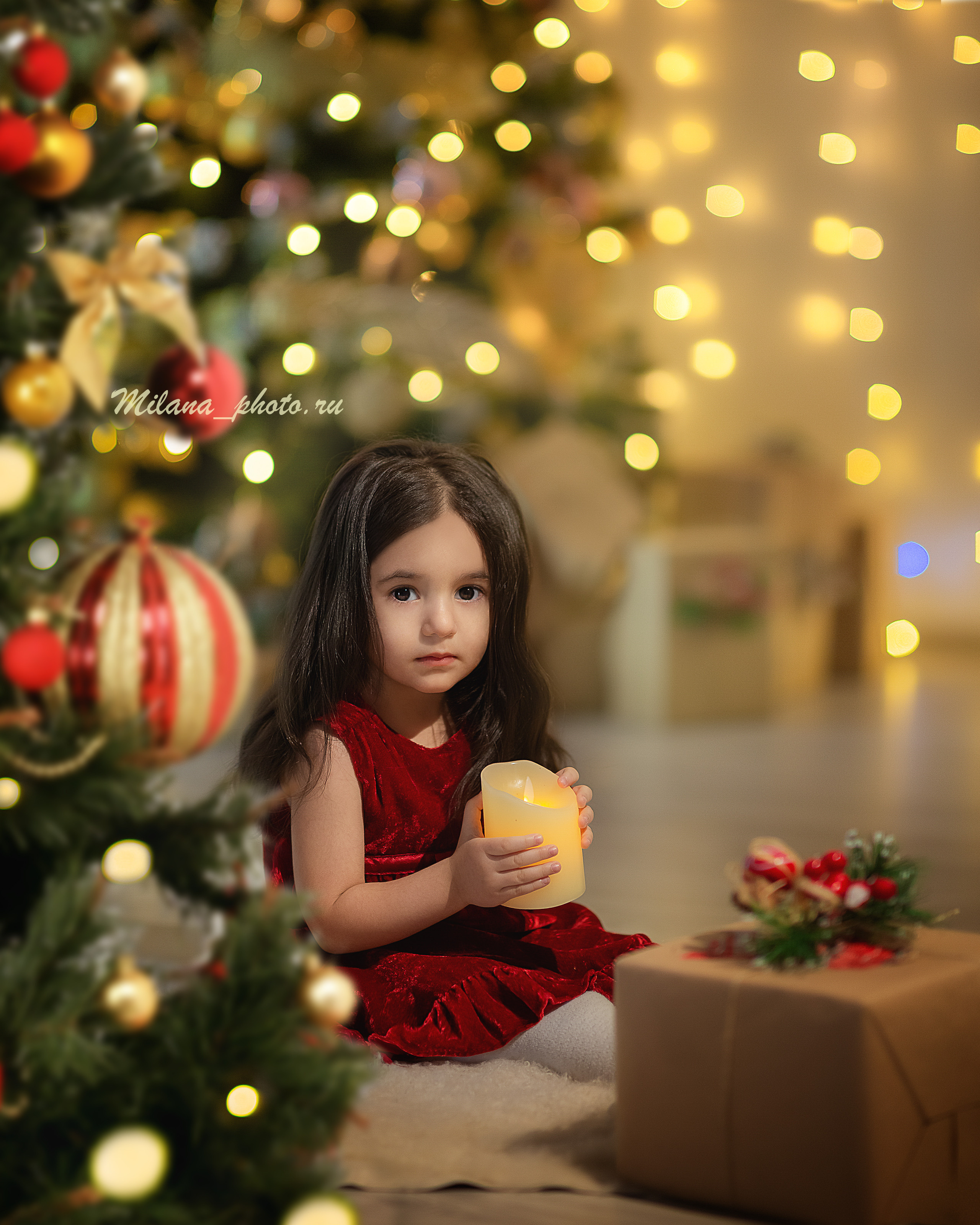 Маленькая девочка  сидит перед новогодней елкой со свечей в руках
