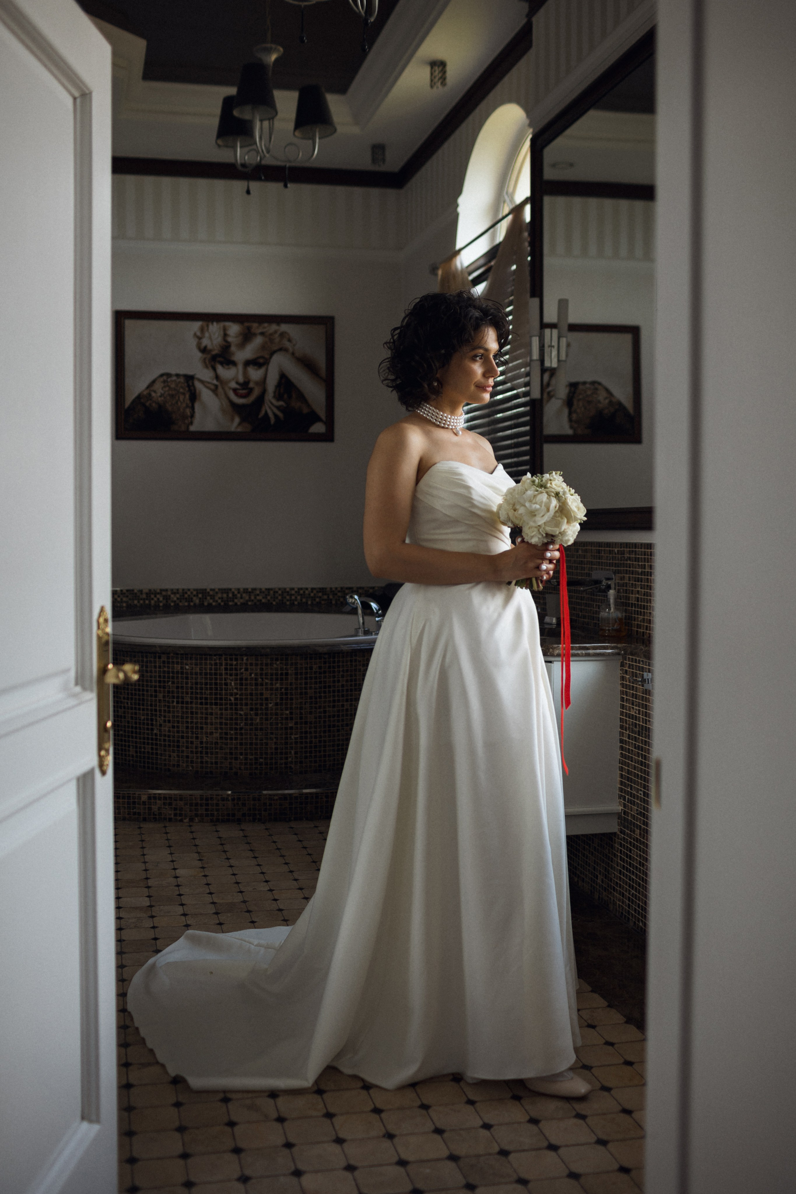 Поэтичный кадр в стиле кино: невеста у окна ванной комнаты Архангельска, свет падает на кружевное платье, а полузакрытая дверь создает границу между приватным и торжественным