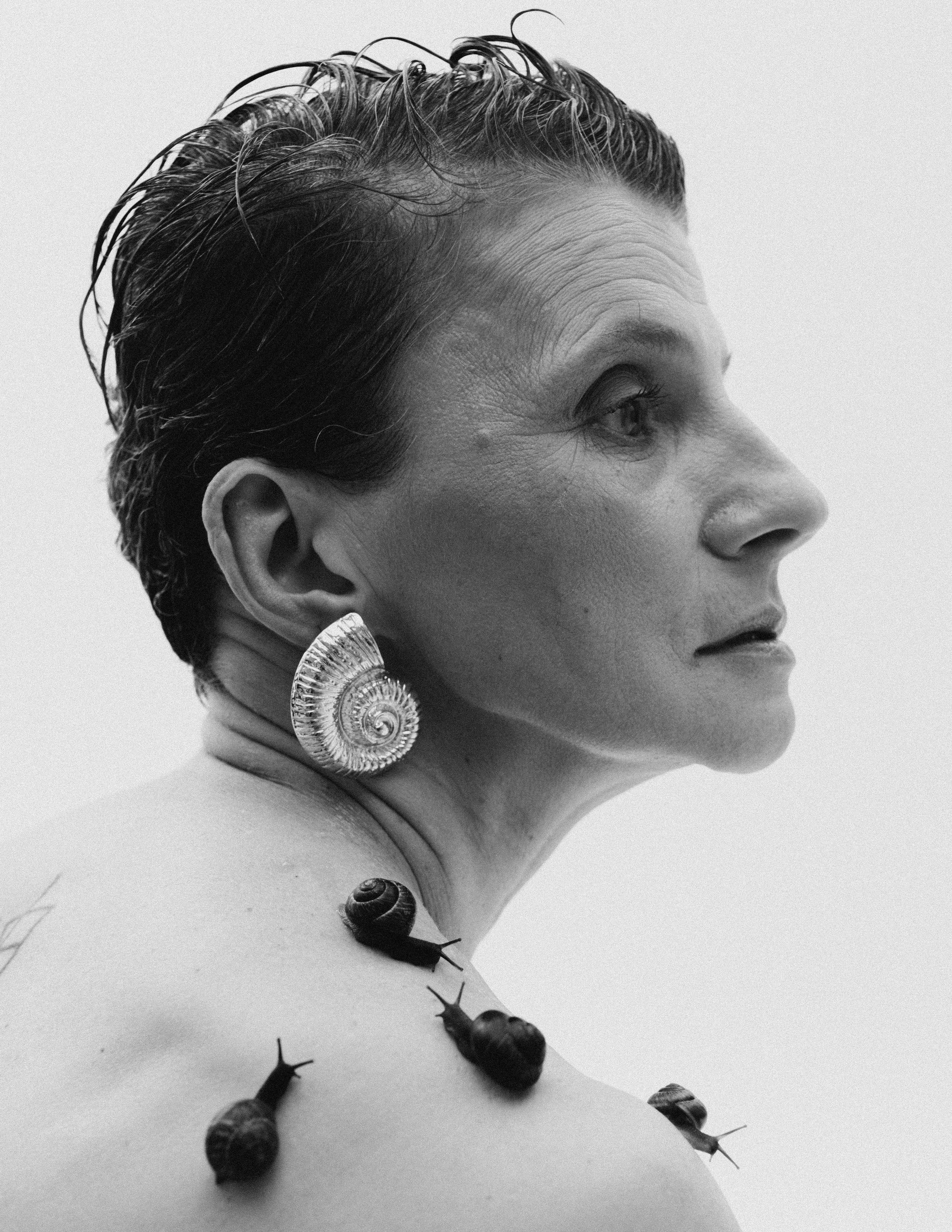 Портрет возрастной модели с улитками на теле и крупной сережкой в форме раковины моллюска.