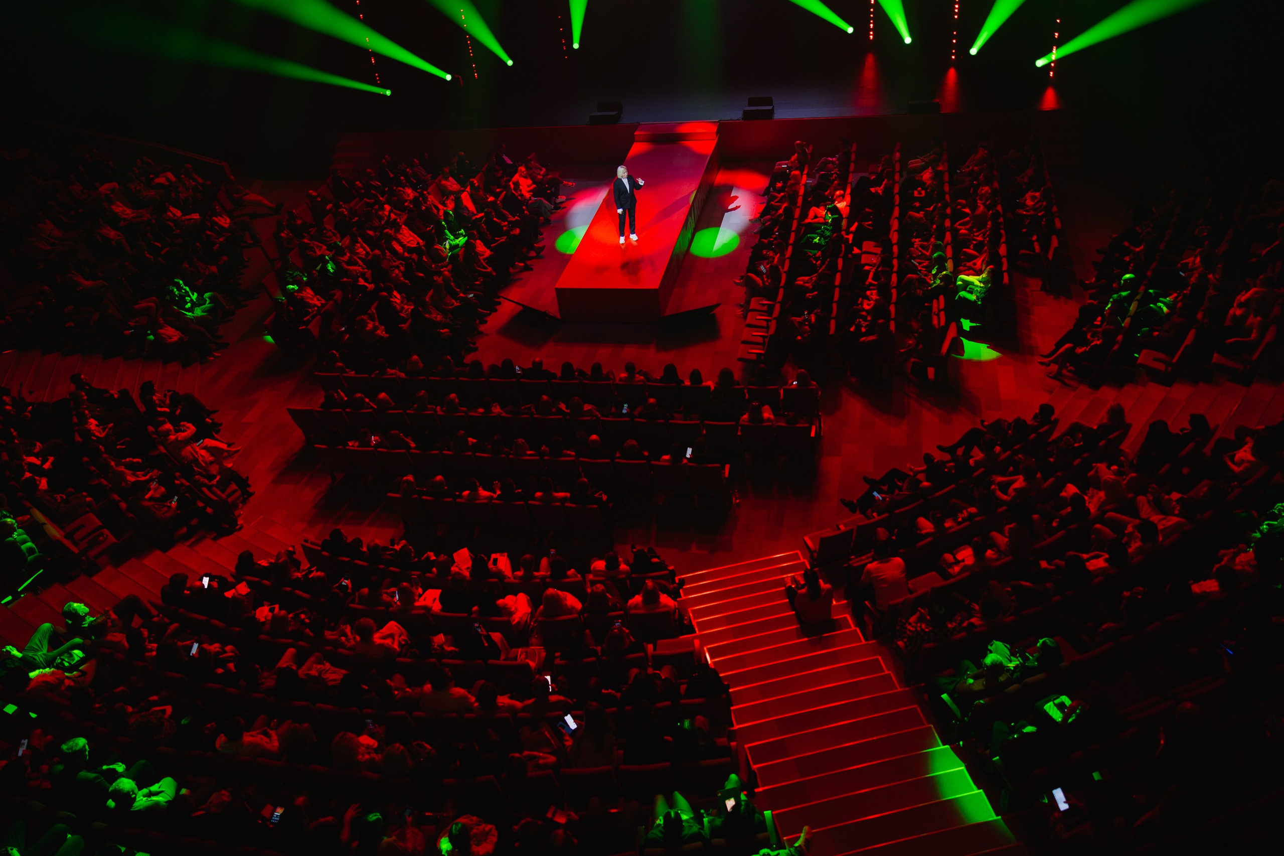 Вид на зал с красной дорожкой: Масштабный план площадки сверху, центральный проход и заполненные ряды зрителей в зеленом свете.