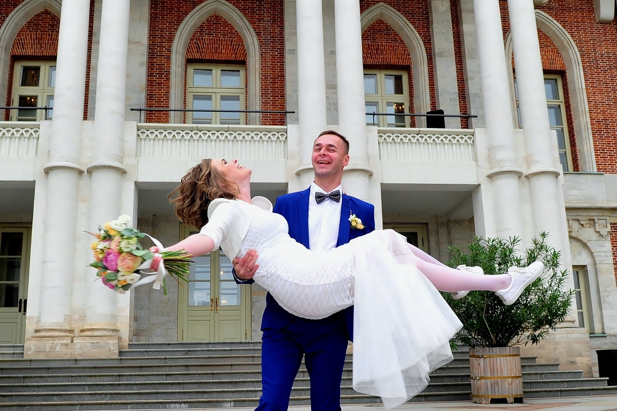 Свадьба в Царицыно хмурым прохладным летним днём, жених несет невесту на руках на фоне дворца из красного кирпича и белого камня, невеста хохочет