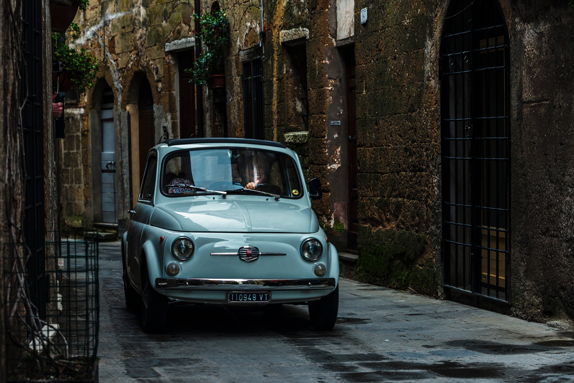 Винтажный голубой Fiat 500 припаркован в узком каменном переулке в Пьенце, Италия, в условиях мягких теней.