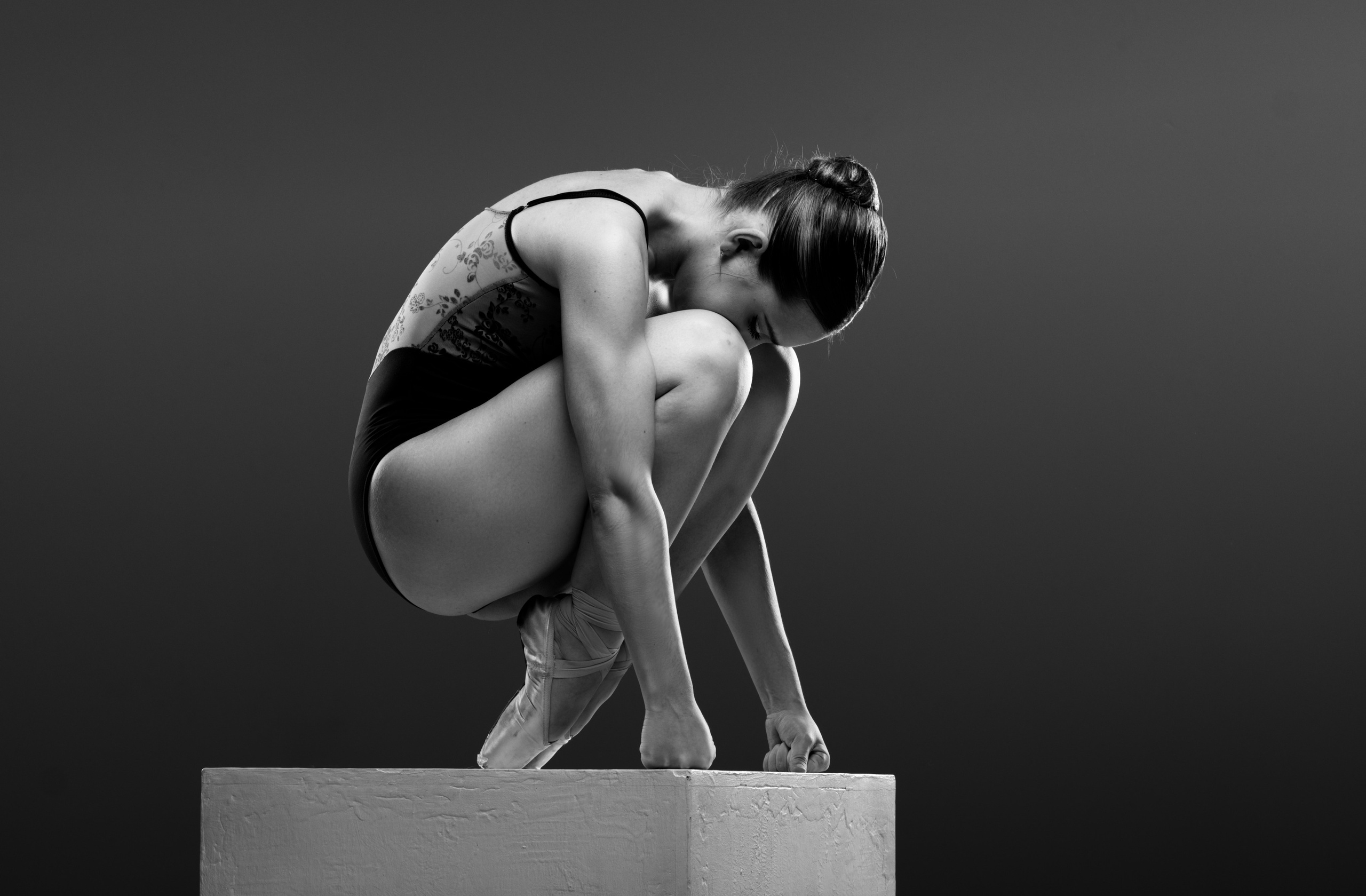 Спортивный фотограф в Тюмени: портфолио балерины для контента в социальных сетях. Черно-белое фото балерины в закрытой скульптурной позе на белом кубе. Акцент на геометрии человеческого тела, лаконичности формы и игре теней.
