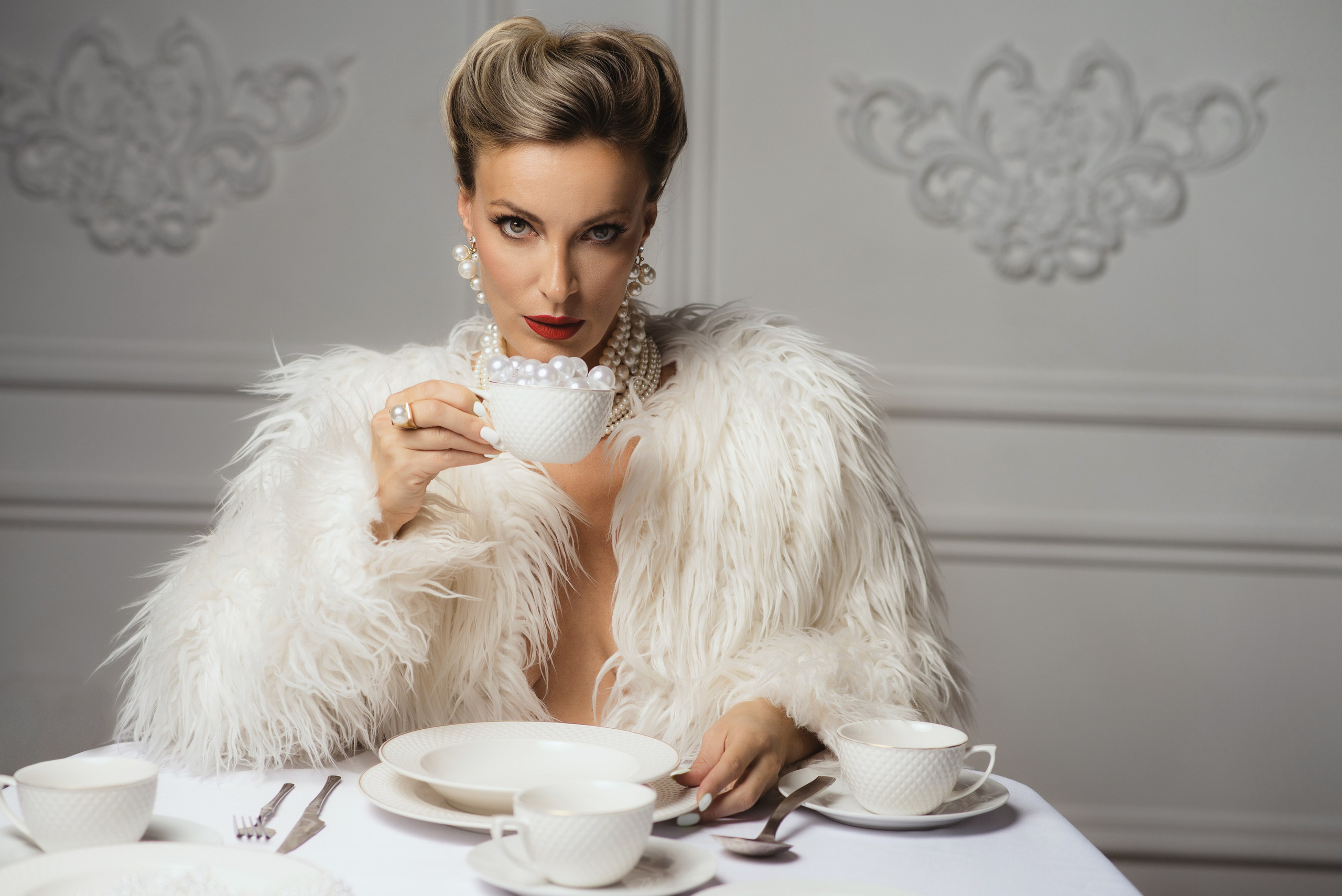 Женщина в белой шубе и жемчужных украшениях держит в руке белую чашку, наполненную крупным жемчугом
