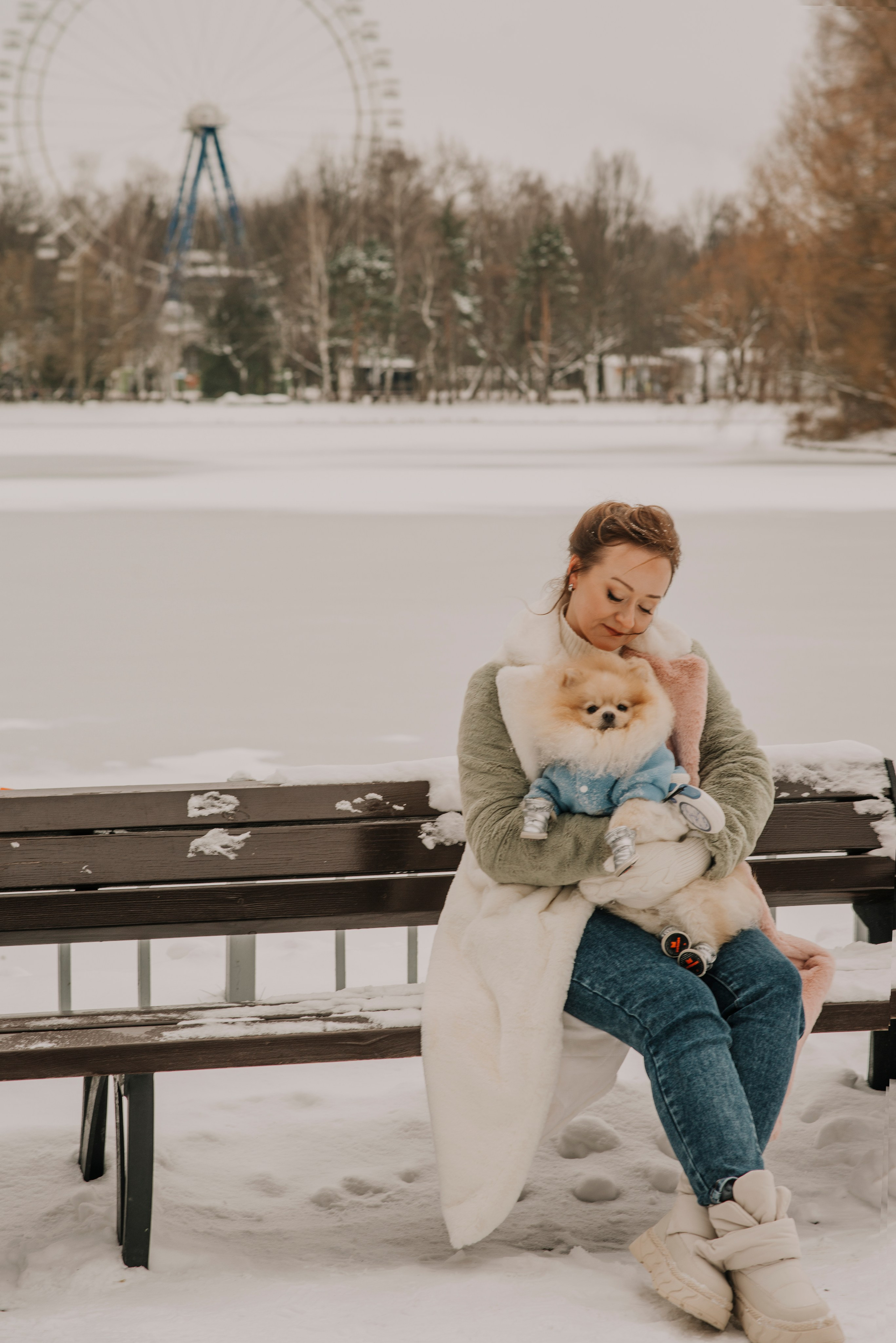 Девушка сидит на скамейке на фоне замерзшего пруда в парке с собакой шпицем на руках