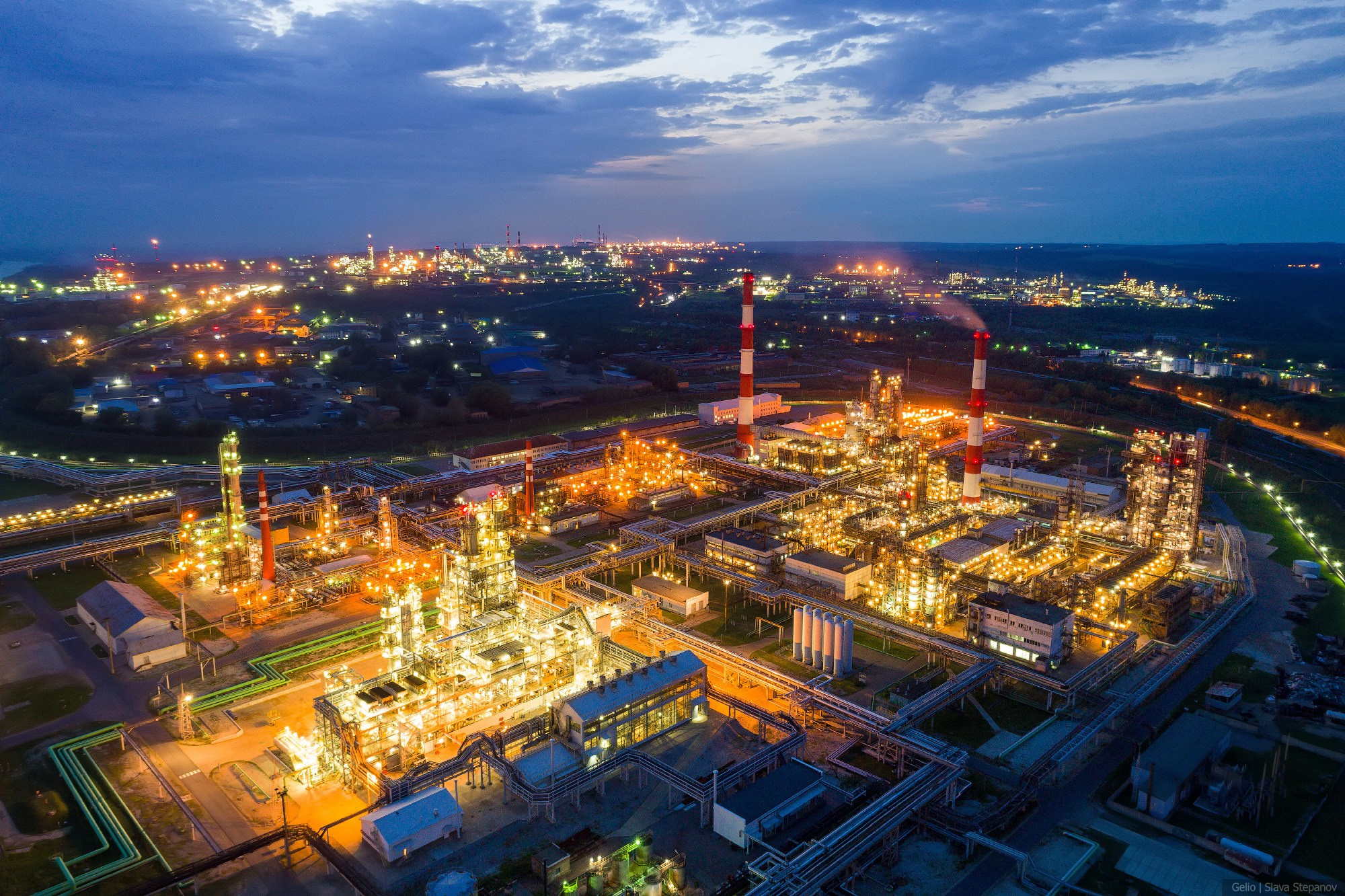 Масштабная панорама нефтеперерабатывающего завода в вечерних сумерках, ярко освещённая индустриальная инфраструктура с высокими трубами, фото Слава Степанов.