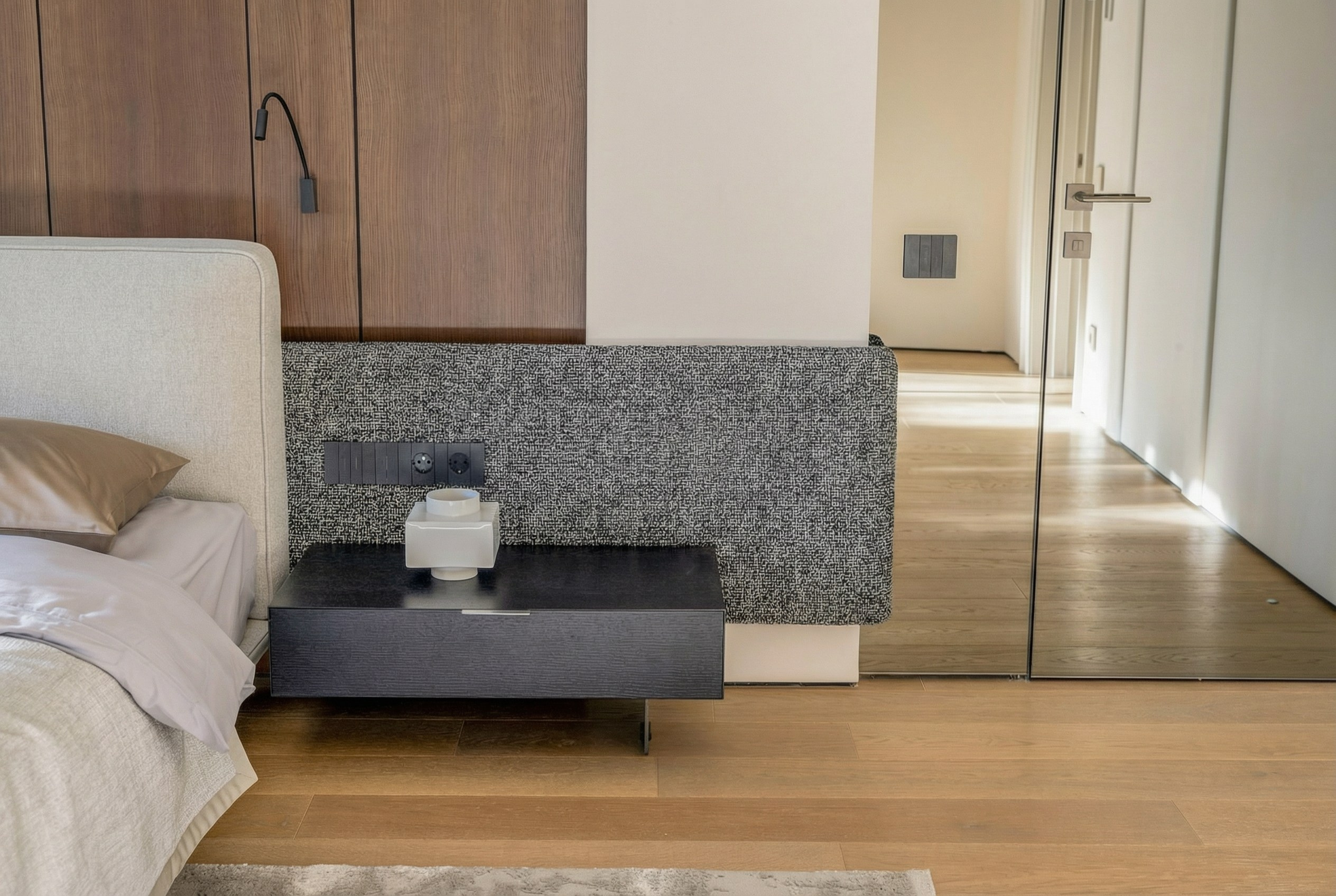 Деталь прикроватной зоны в мастер-спальне: подвесная черная тумба, мягкое настенное панно из рогожки и шпонированные панели, проект Валерии Котович.