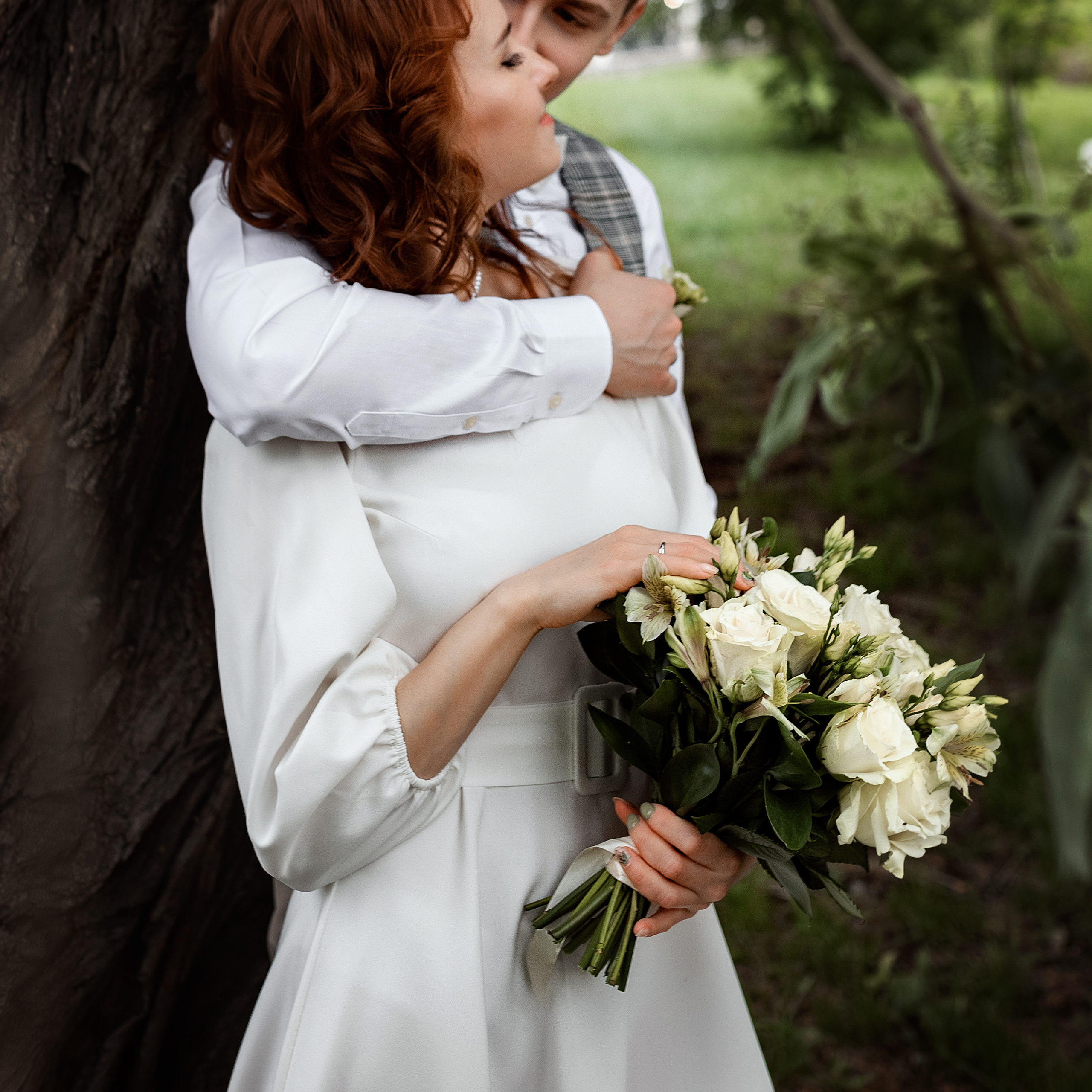 Молодожены на свадебной фото прогулке целуются у широкого ствола дерева
