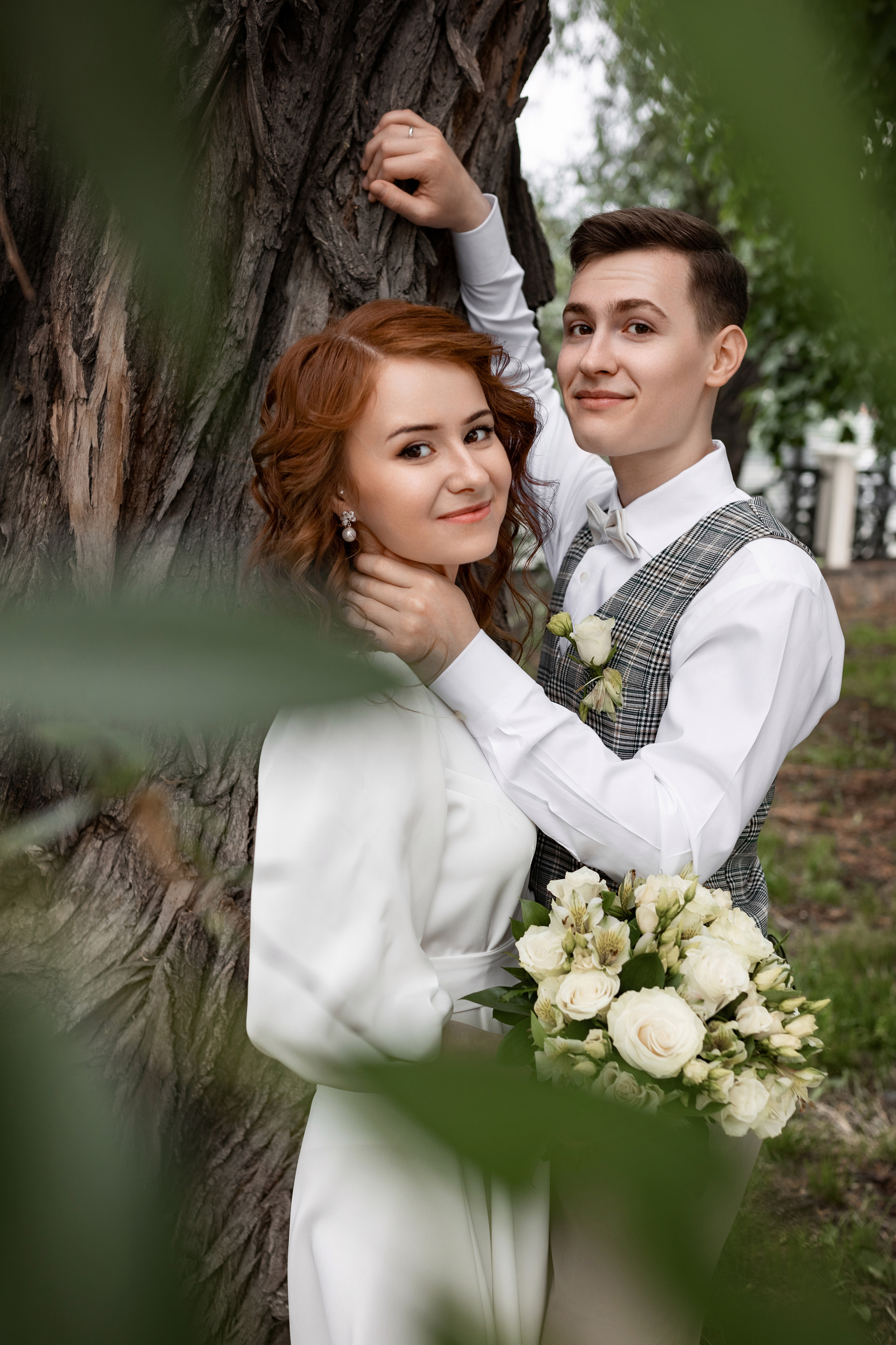 Молодожены на свадебной фото прогулке целуются у широкого ствола дерева