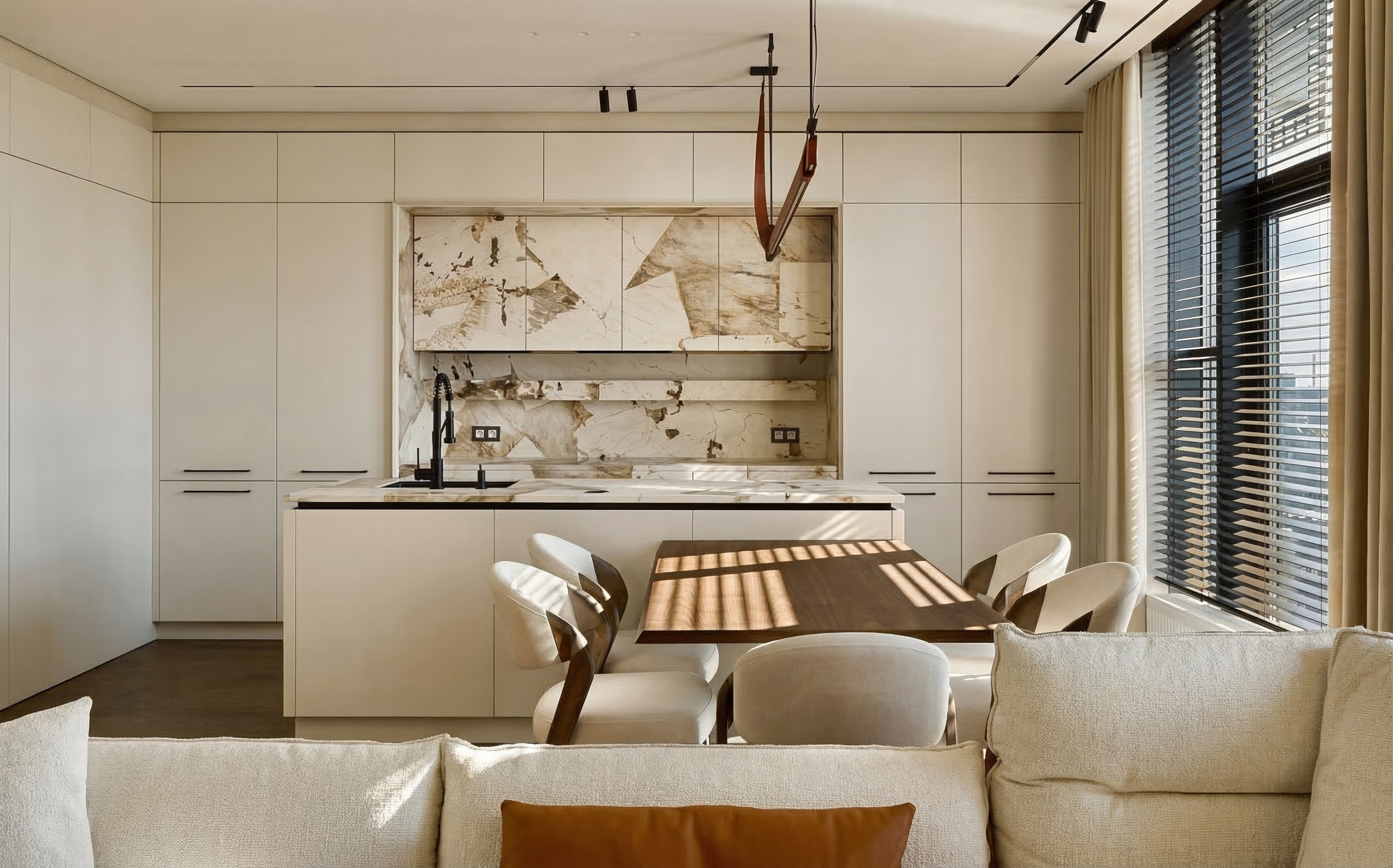 Современная светлая кухня с островом, фартуком из натурального камня с крупными прожилками и дизайнерским линейным светильником на кожаных подвесах.