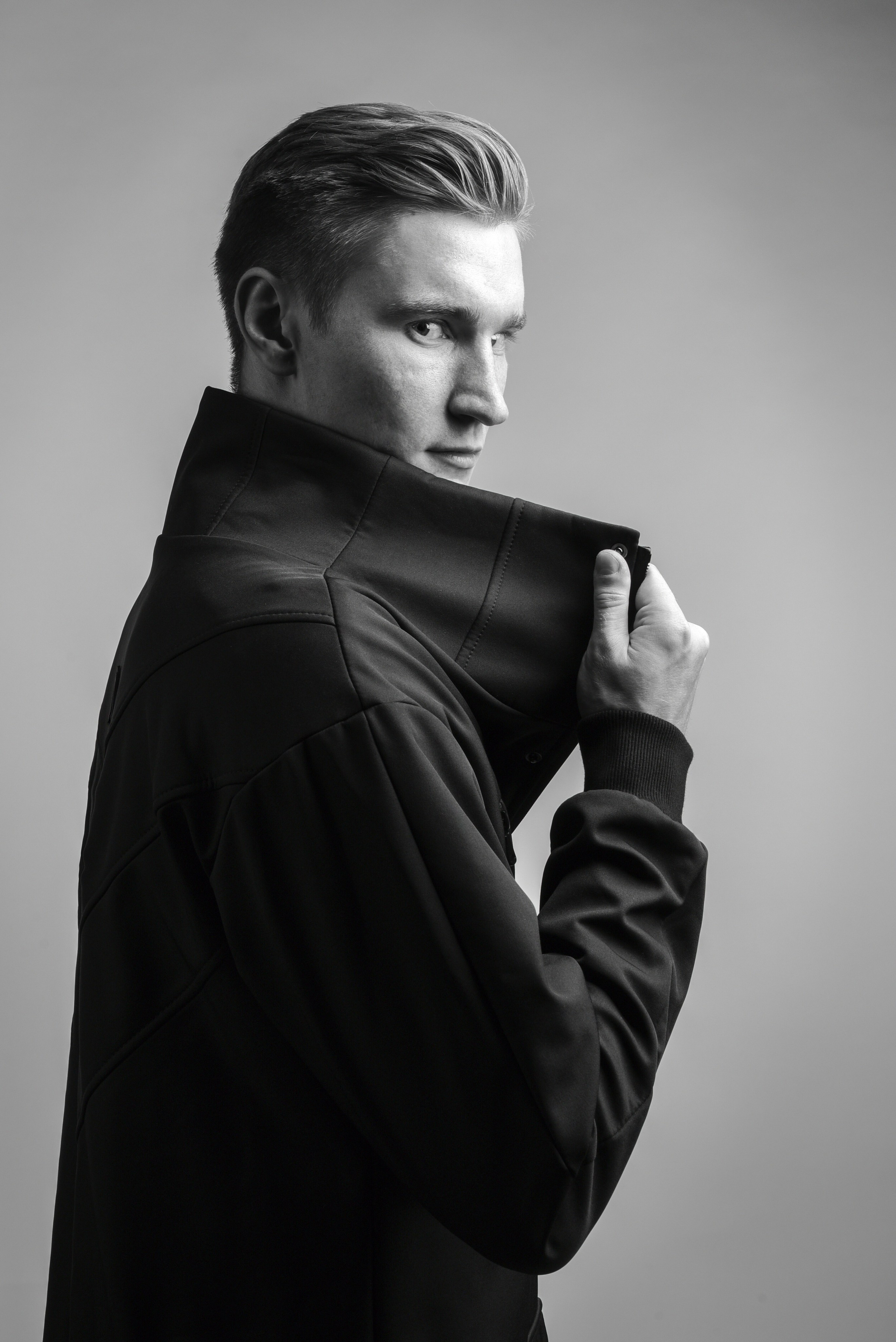 Молодой человек в чёрном плаще на белом фоне. Стоит боком. Закрывает часть лица закрыта широким воротником.