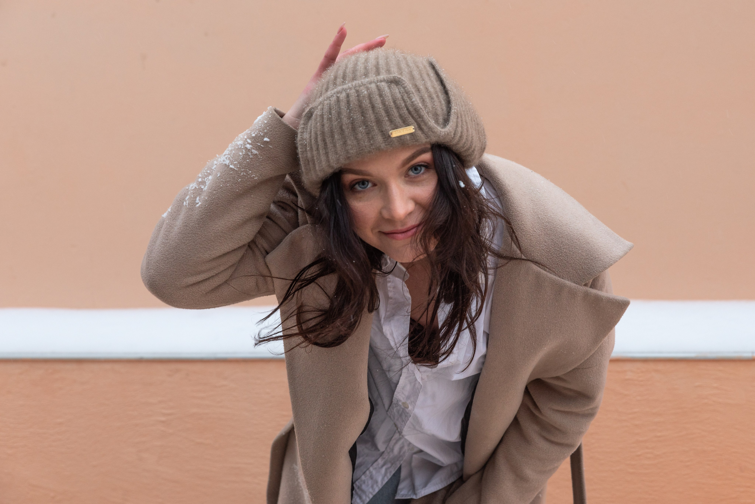 Крупный план. Женщина в бежевом пальто и вязаной шапке наклони наклонилась к камере и улыбается, одной рукой придерживает шапку. На заднем плане бежевая стена.