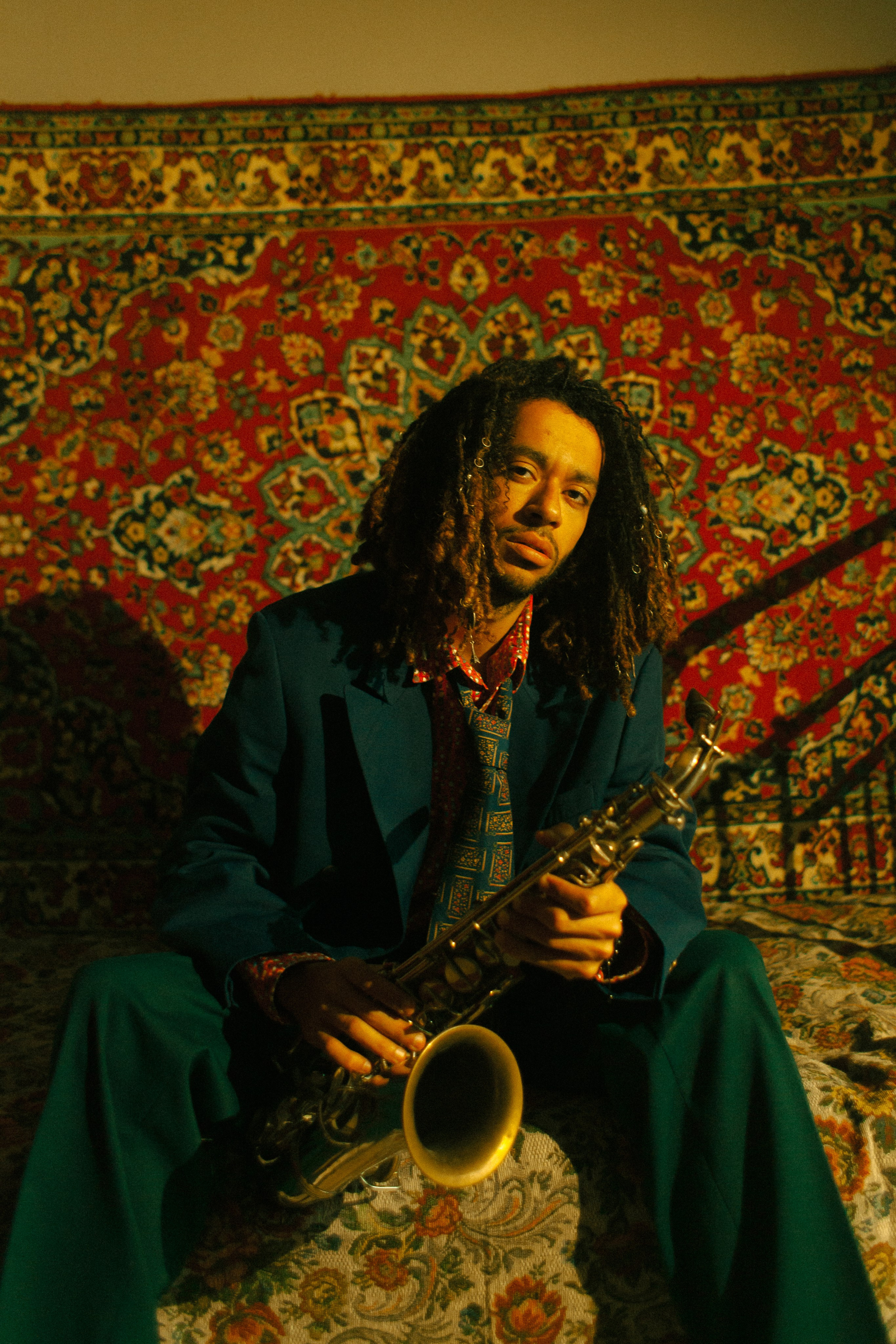 Темнокожий парень в зеленом пиджаке и саксофоном в руках сидит на фоне советского ковра