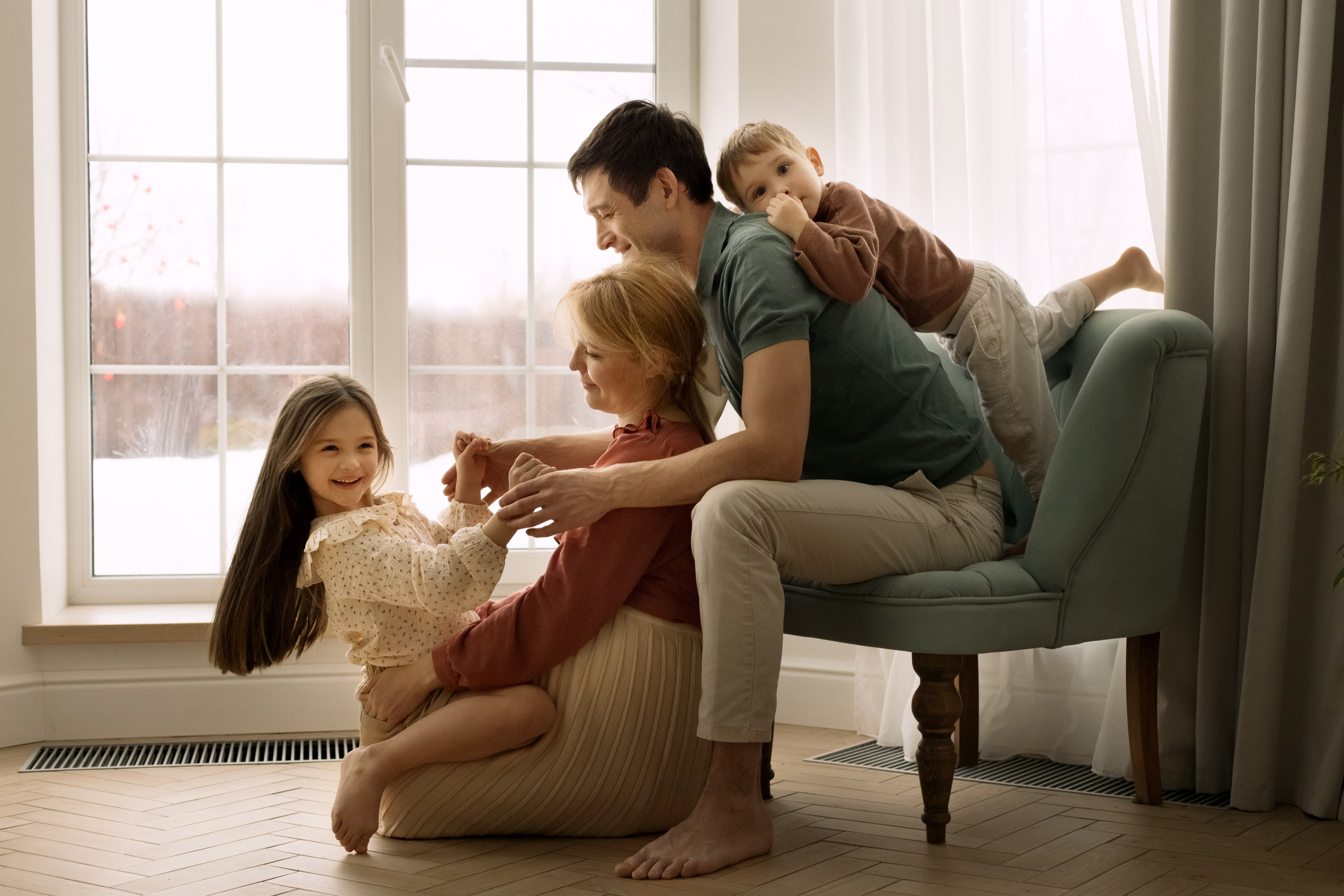 Беременная мама, папа, дочка и сын обнимаются, сидя на кресле у большого окна.