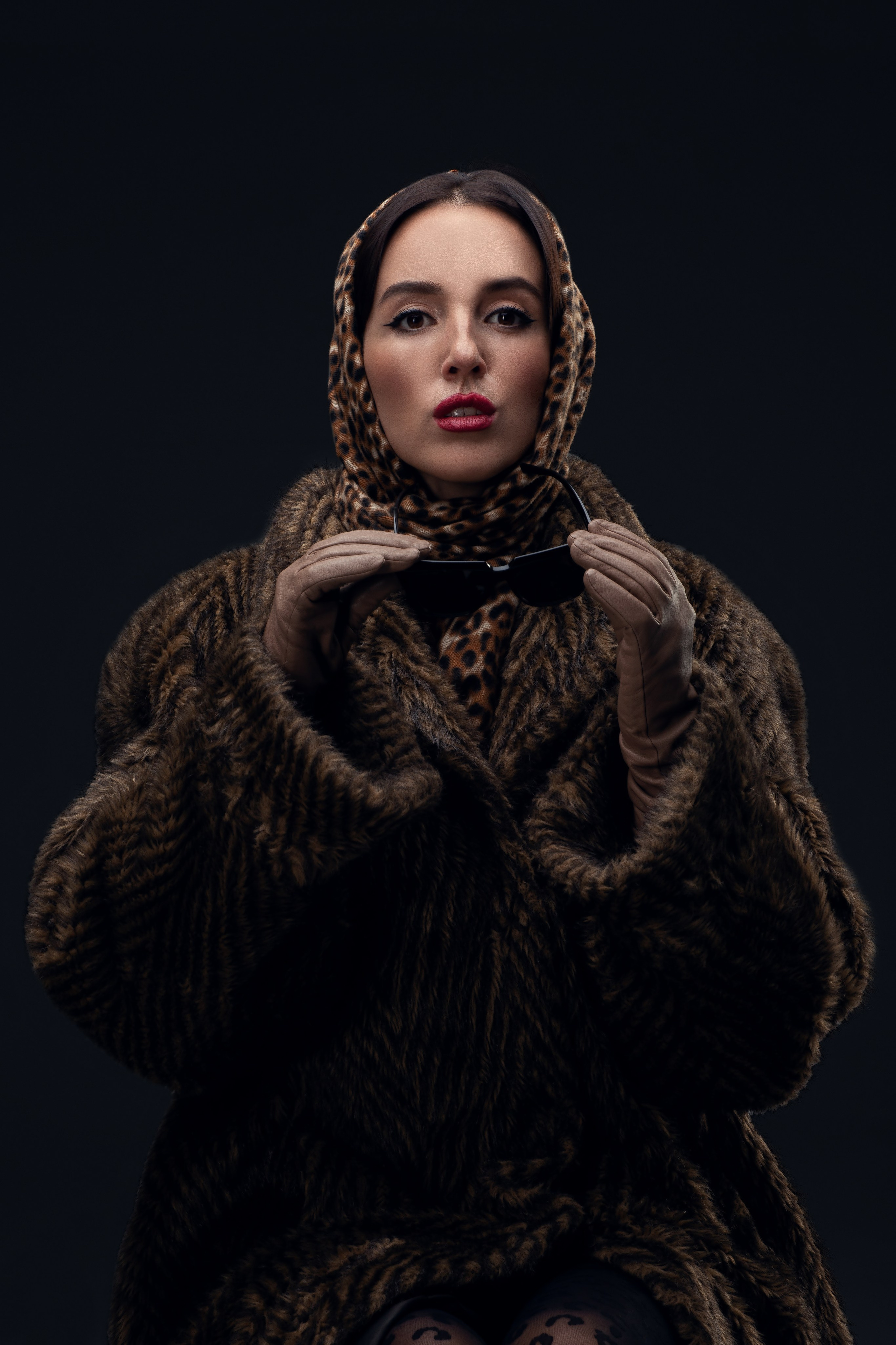 Стильный портрет женщины в леопардовом платке и меховой шубе, держащей солнцезащитные очки. Элегантный образ с ярким макияжем. Гламурный портрет: женщина в стиле ретро, сочетающем животный принт и роскошную меховую шубу. Фотография передает атмосферу шика и уверенности.