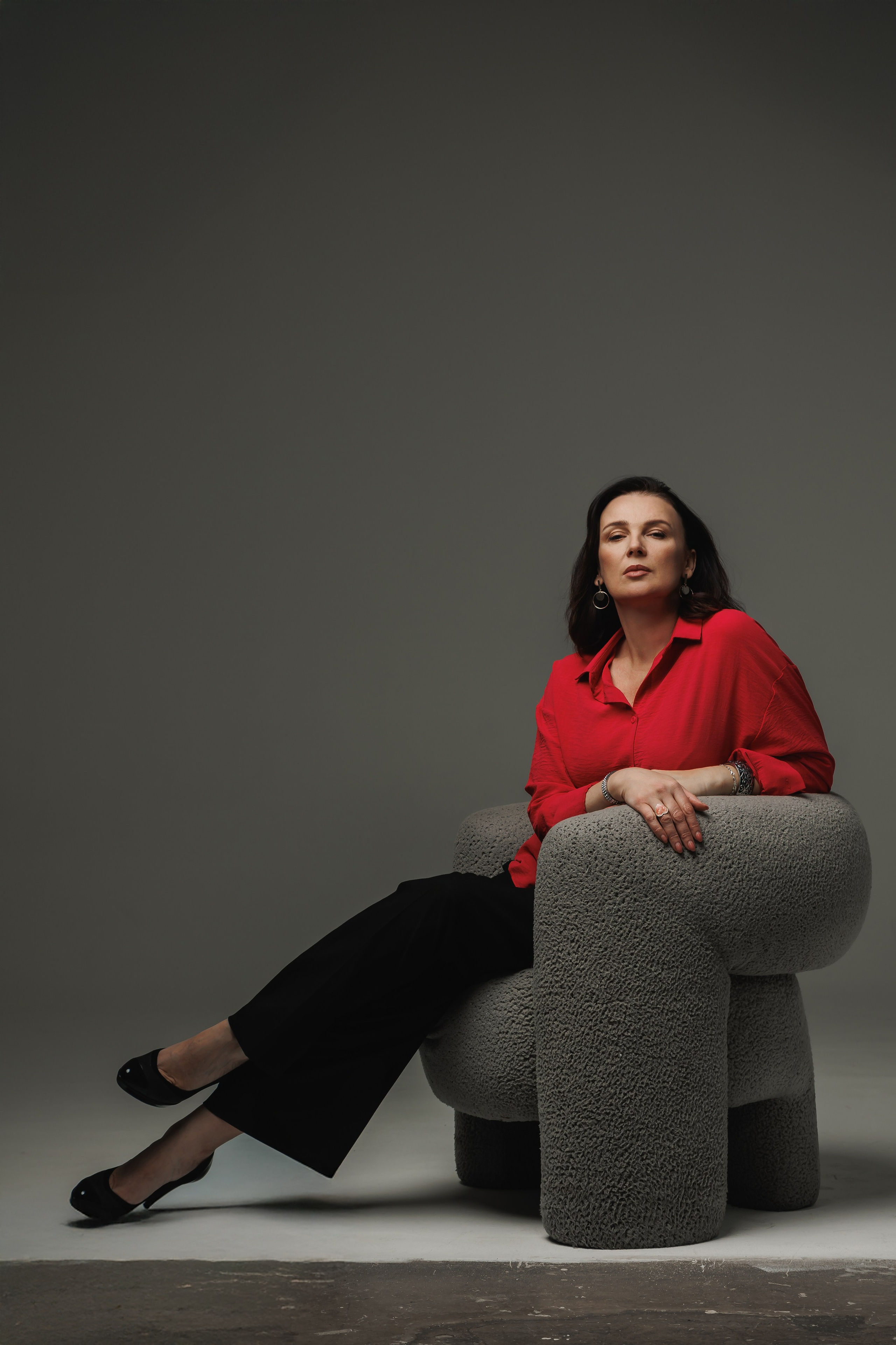 Психолог в позе консультанта, сидя в кресле на однотонном сером фоне. Спокойный и уверенный образ.