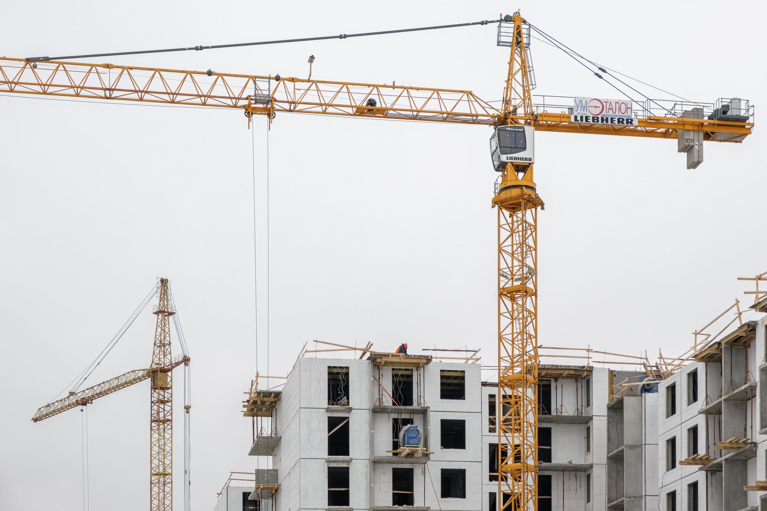 На изображении видно два жёлтых крана, установленных на строительной площадке. Один кран находится ближе к камере, второй расположен чуть дальше. На заднем плане видны строящиеся здания с бетонными конструкциями и частично установленными окнами.
