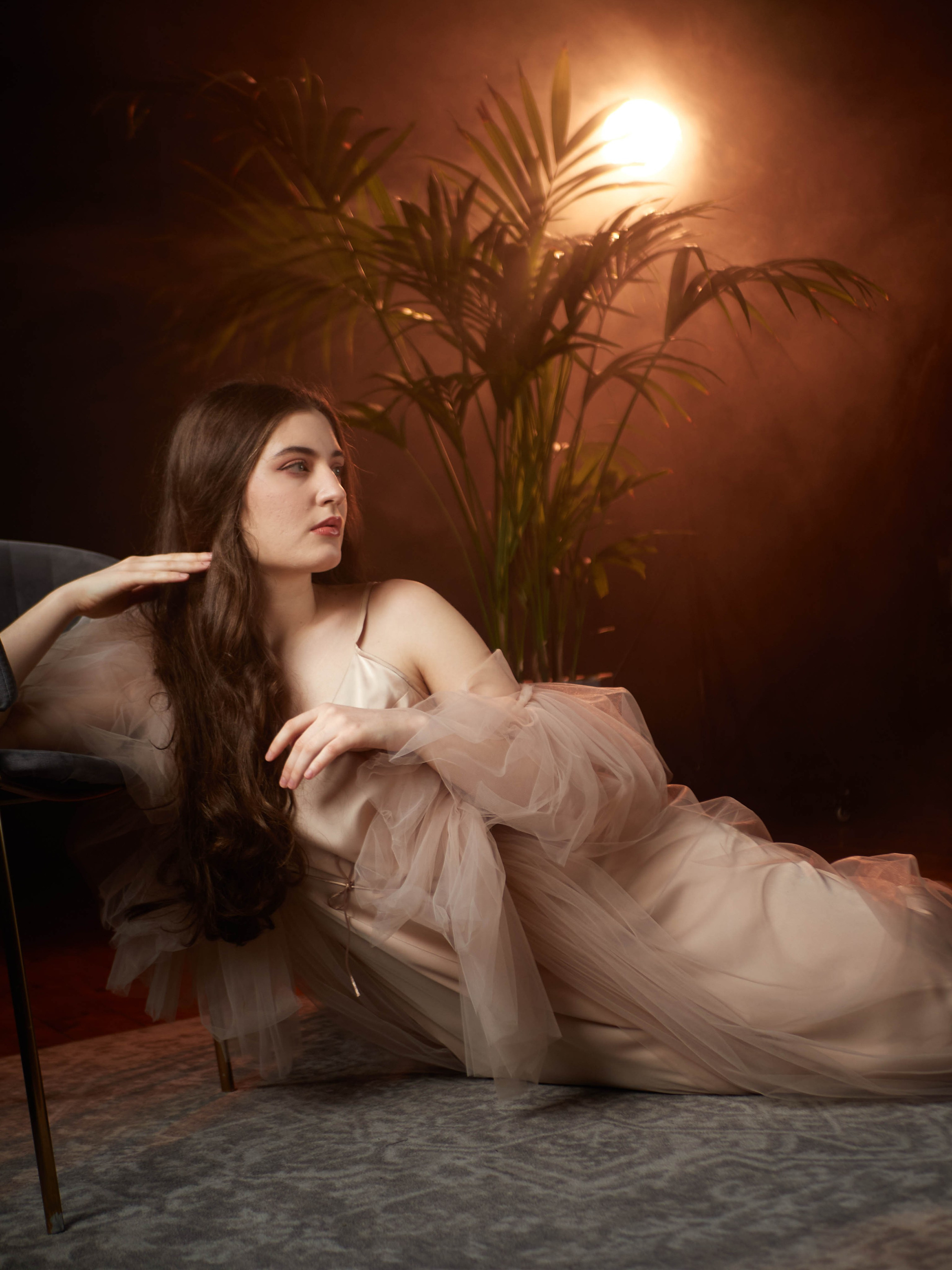 Девушка сидит на полу в бежевом платье на фоне постоянного источника света.