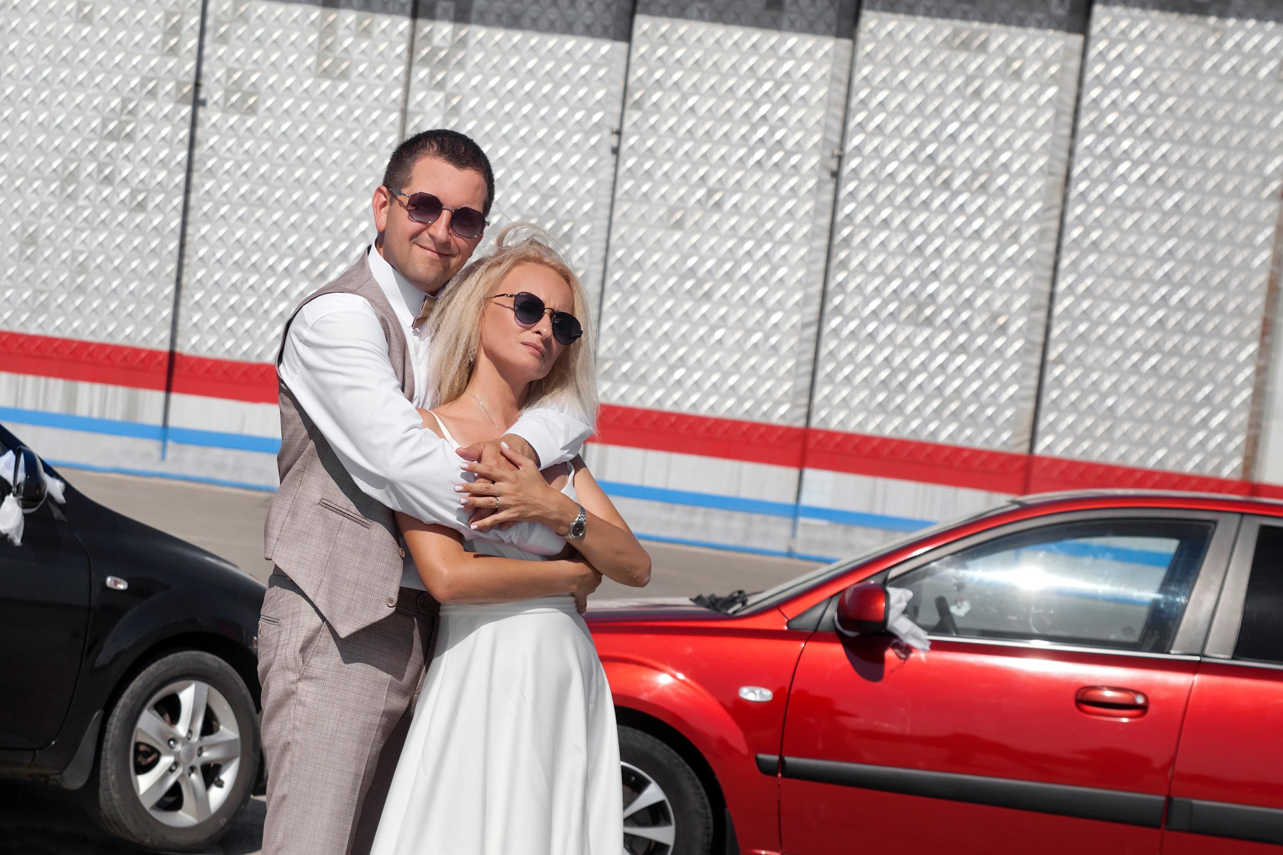 Жених обнимает невесту, позади стоят две машины красного и черного цвета