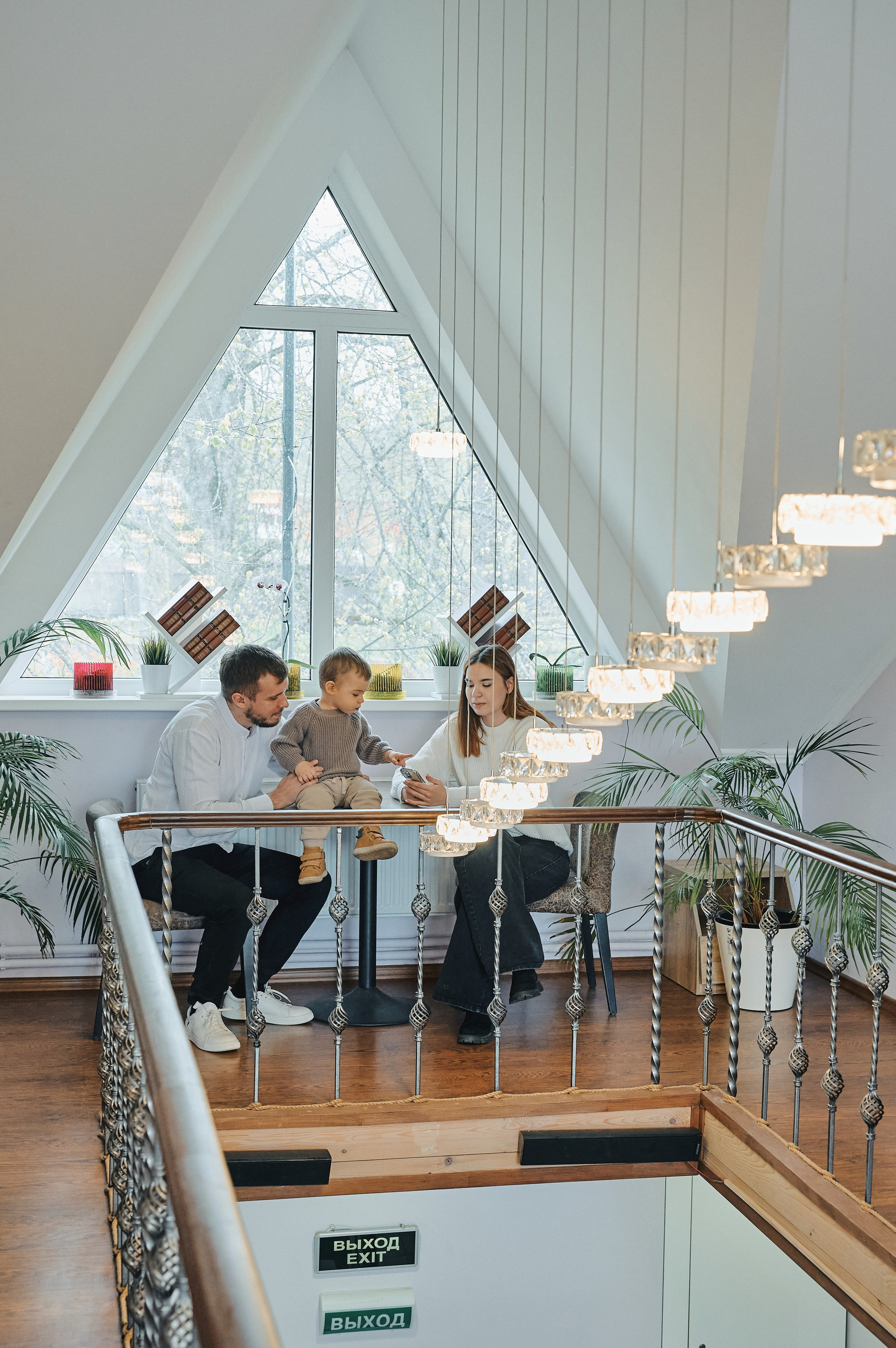 Семья с ребёнком сидит у большого окна на втором этаже, вокруг интерьер с растениями и лампами