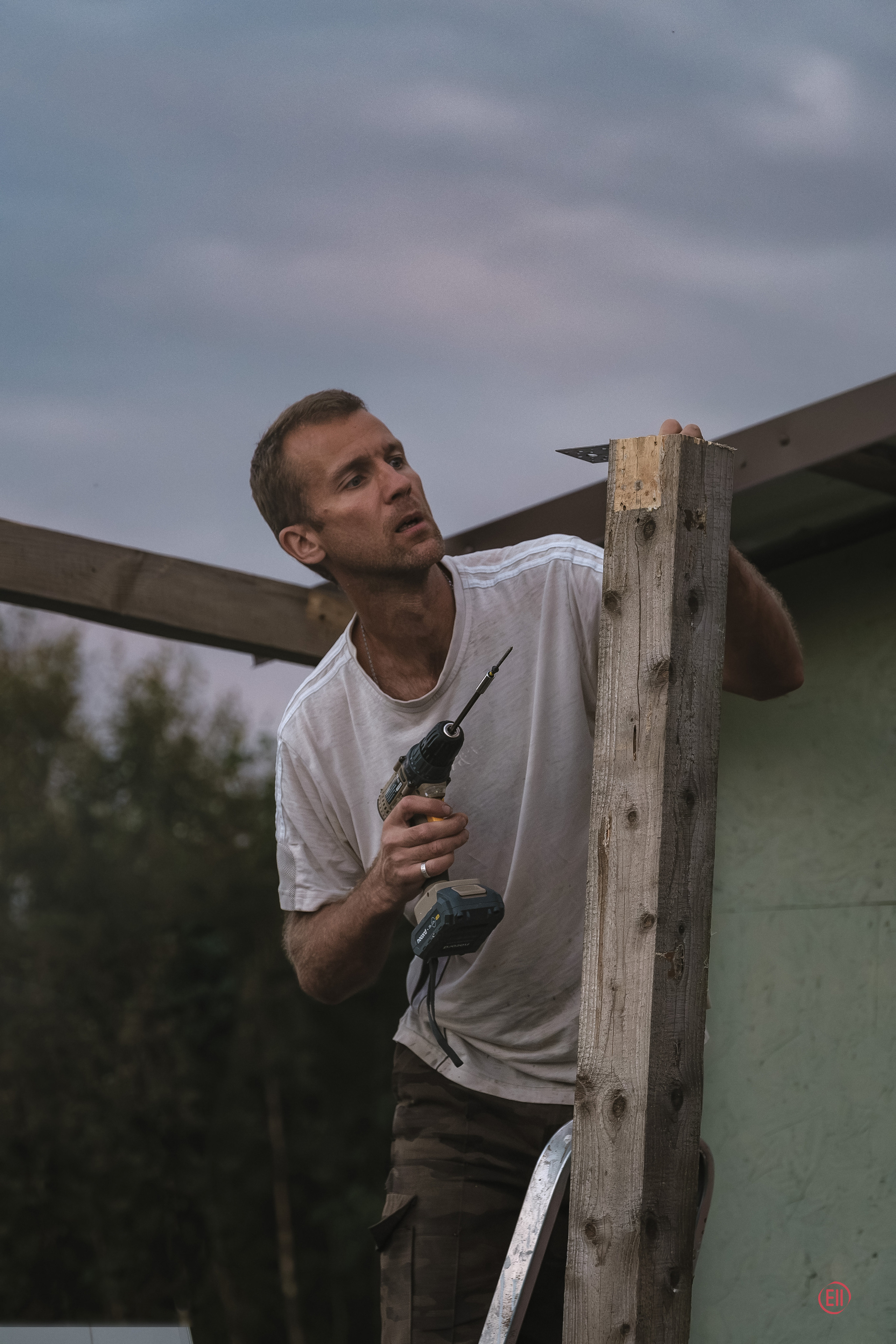 Вечер на даче. Мужчина с дрелью на стремянке пытается прикрутить железный уголок.