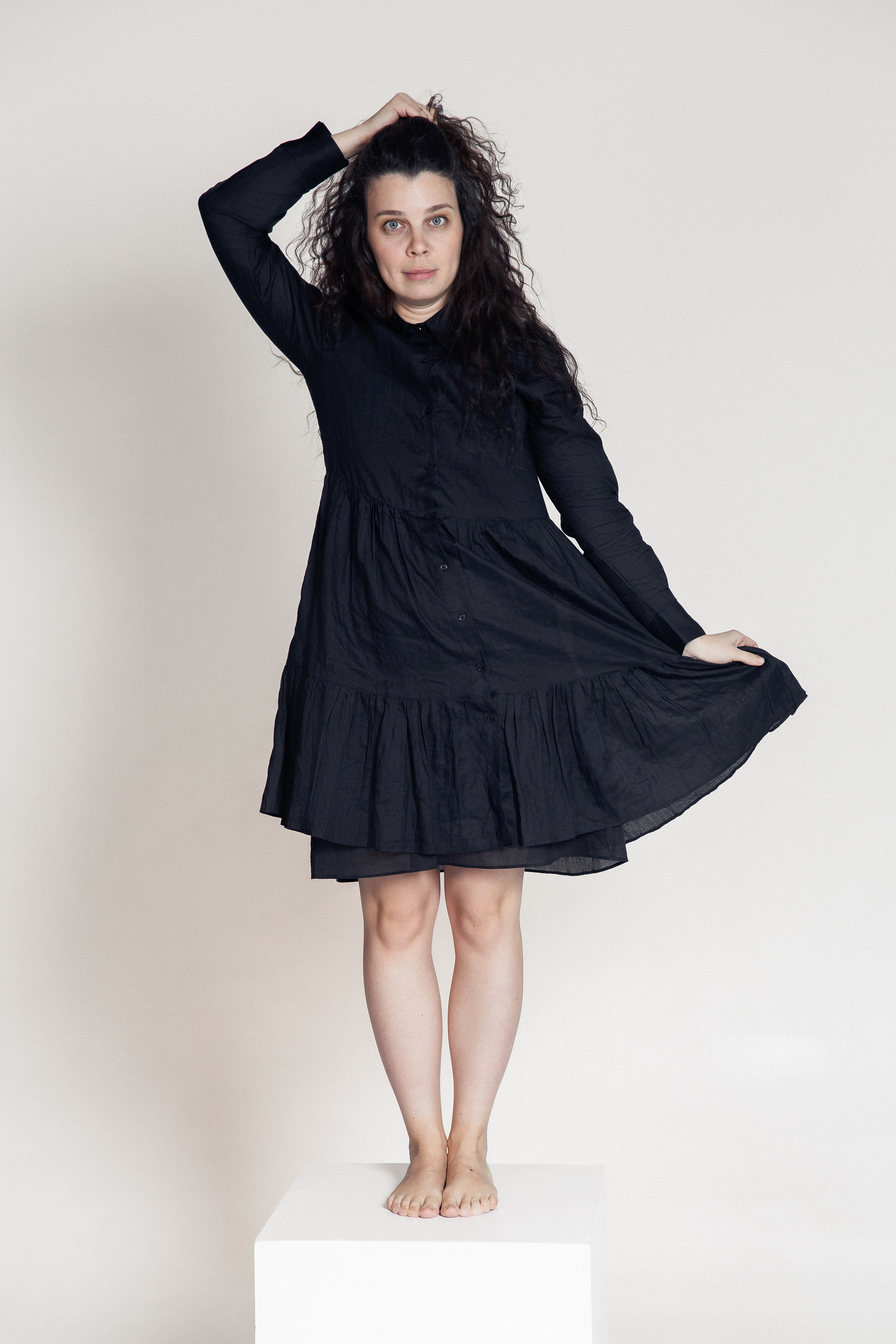 Женщина в чёрном платье стоит на белом кубе, приподняв подол юбки, смотрит в камеру с лёгкой улыбкой.