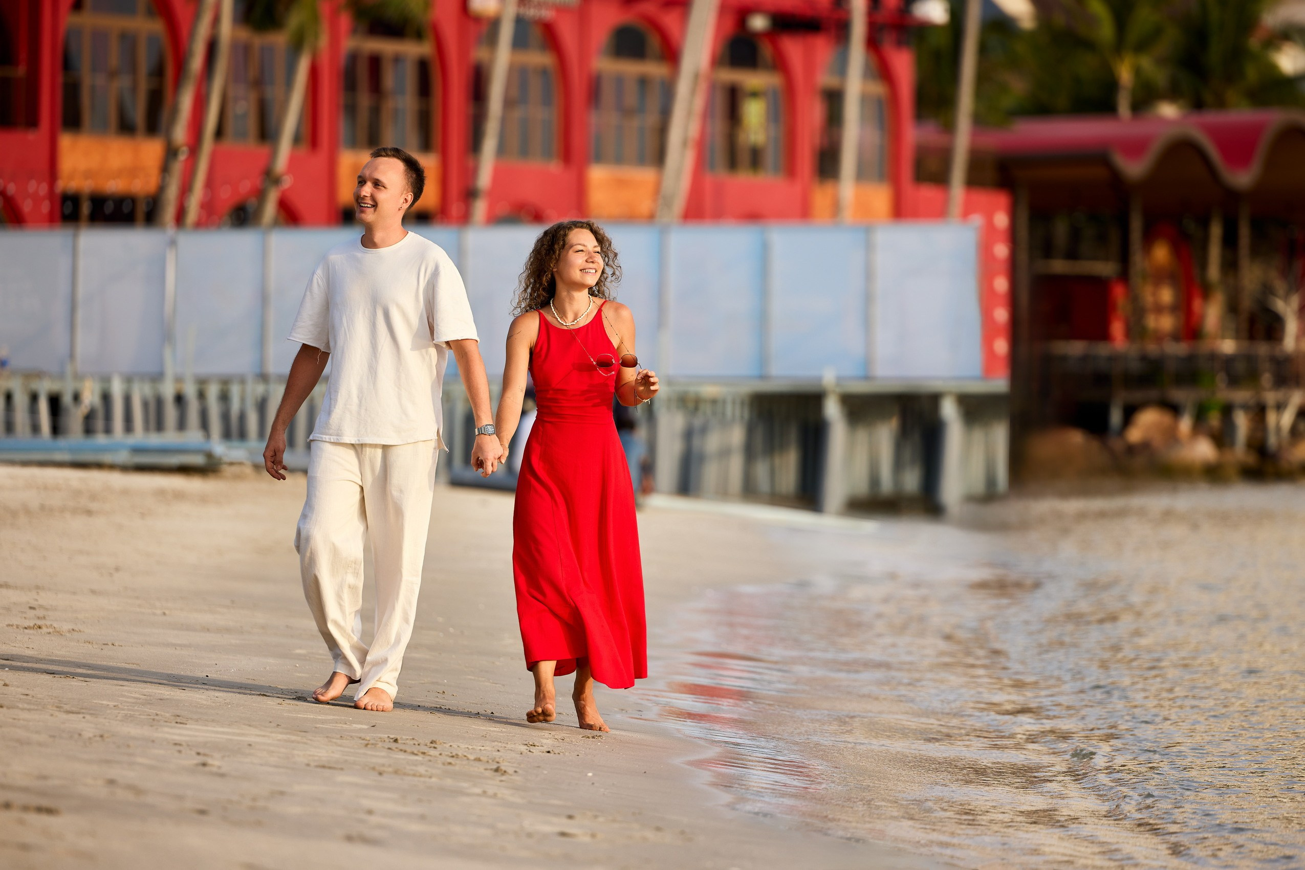 Couple walking hand‑in‑hand along the water’s edge on a sunny sandy beach during a couples photoshoot, captured by the on‑site photographer; red arched building and palm trees in the background, Sunset Town, Phu Quoc, Vietnam.