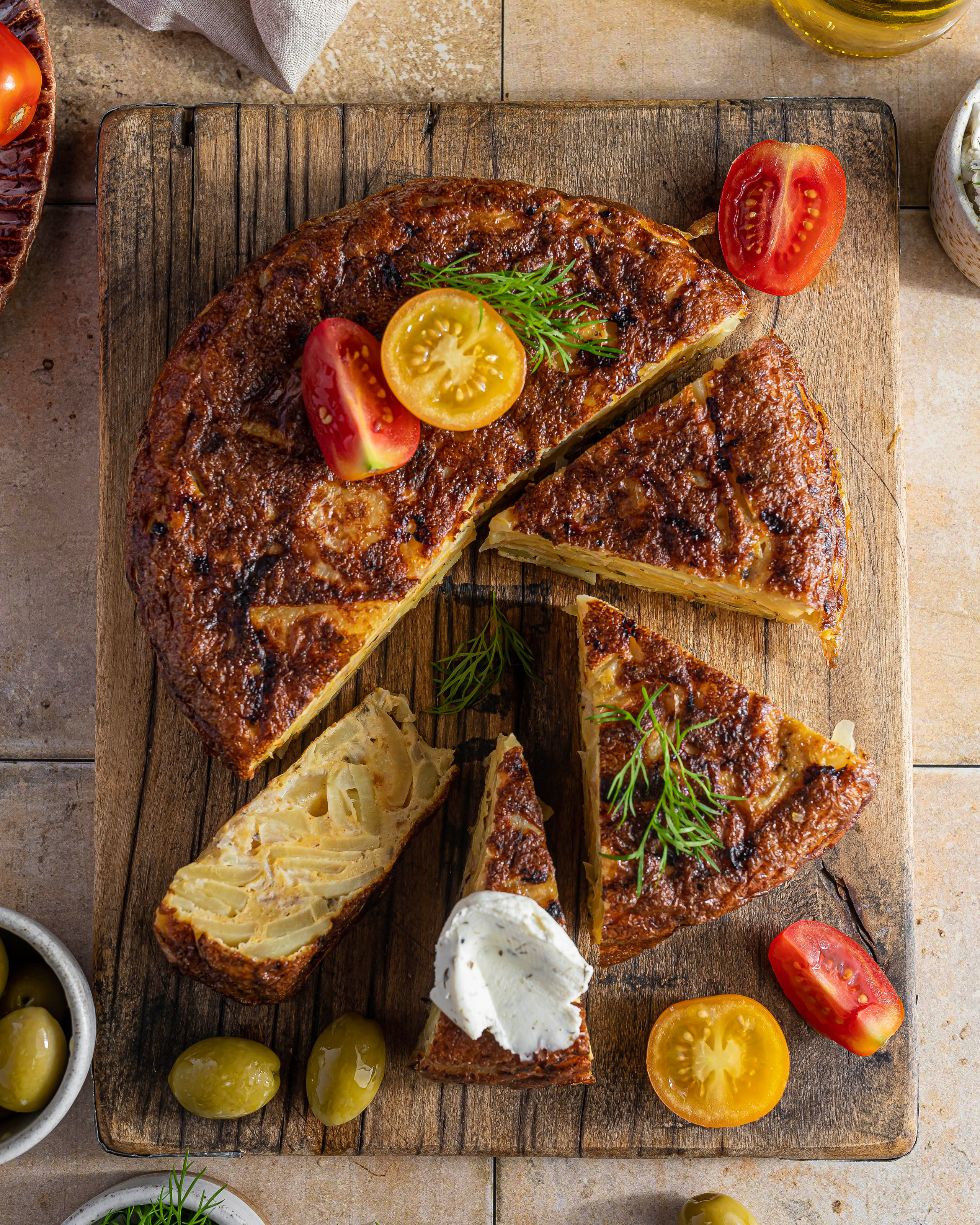 Испанская тортилья с картофелем, украшенная сливочным сыром и свежими томатами, лежит на деревянной доске