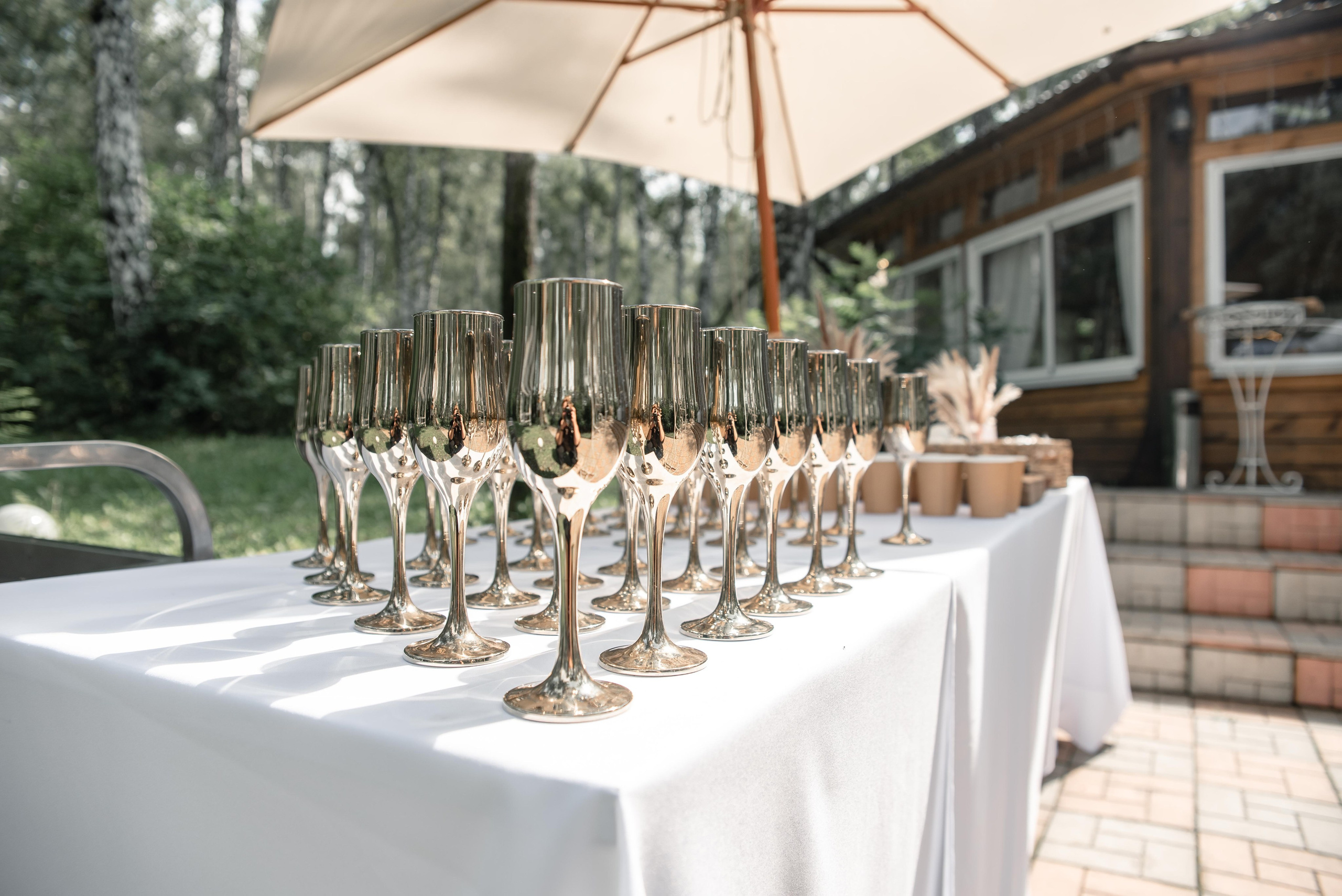 На фото красиво сервированный стол с золотыми бокалами для шампанского, стоящими на белой скатерти. Вокруг видны зеленые деревья и уютный деревянный домик, создающие атмосферу праздника и уюта. Идеальное место для торжественного события! 🥂✨