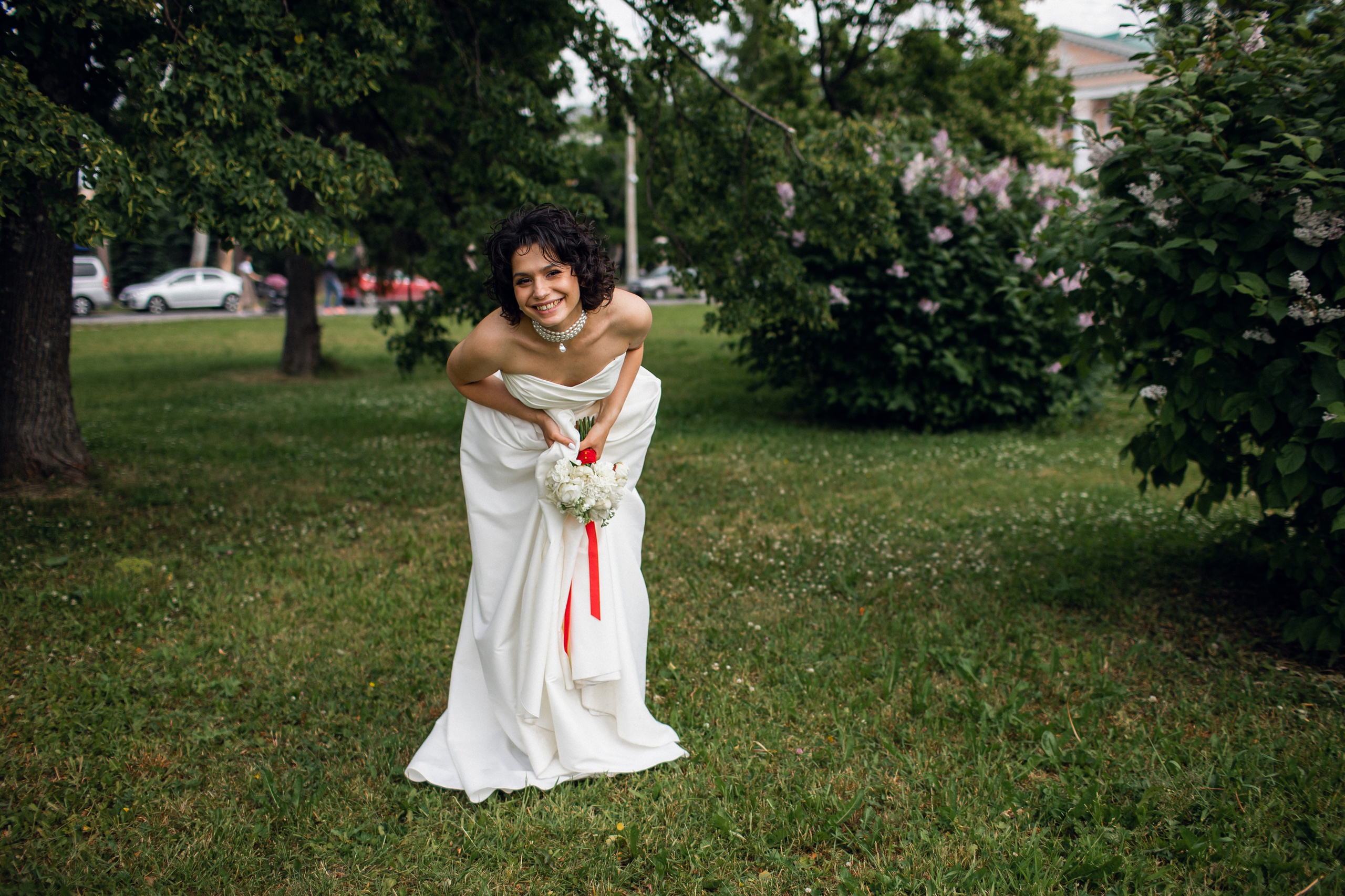 Свадебная фотосессия в стиле lifestyle: невеста с улыбкой кривляется у кустов сирени в Центральном парке Архангельска в летних лучах солнца