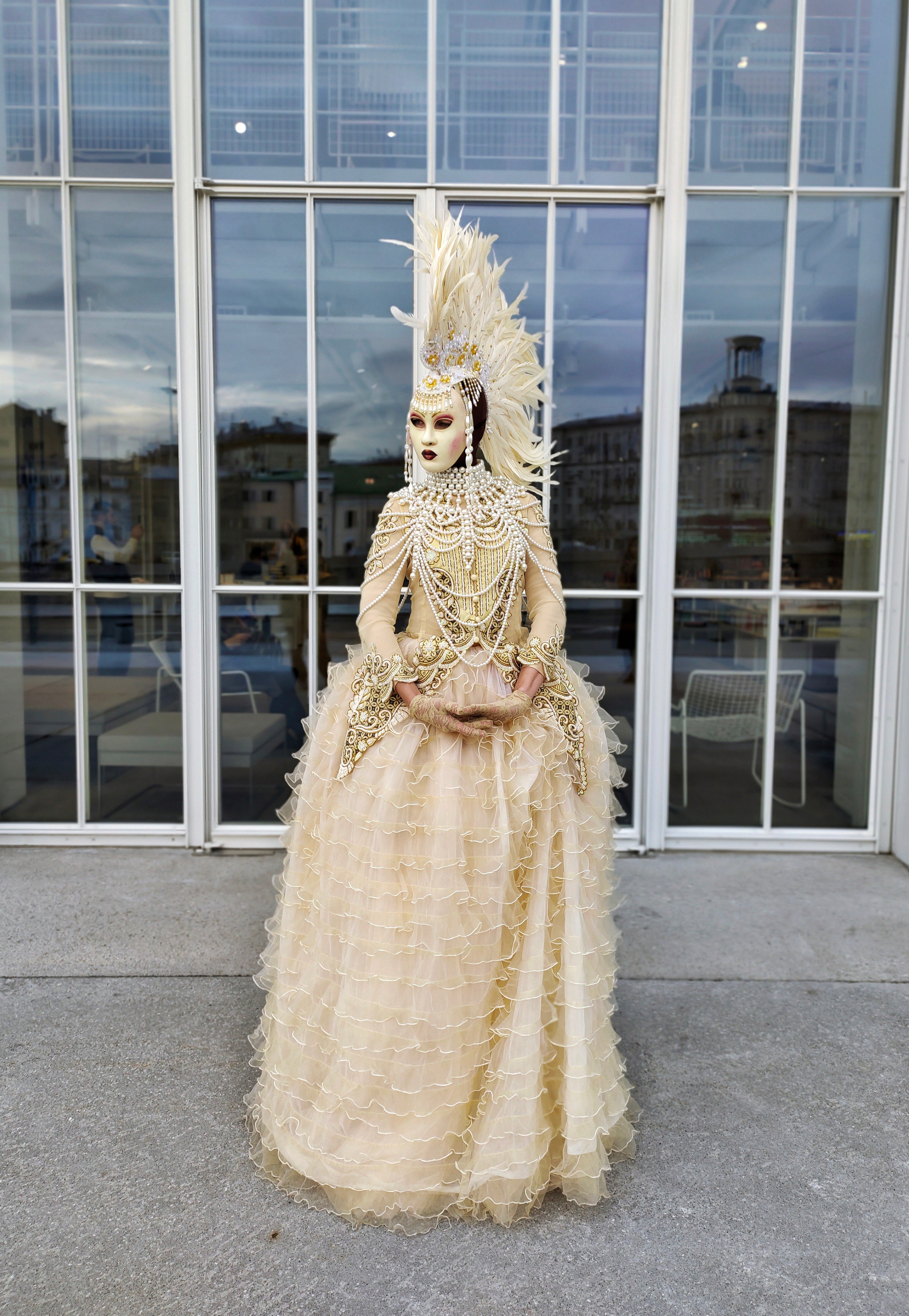 Мим в бежевом платье расшиты стразами в маске и с ирокезом приветствует гостей выставки.