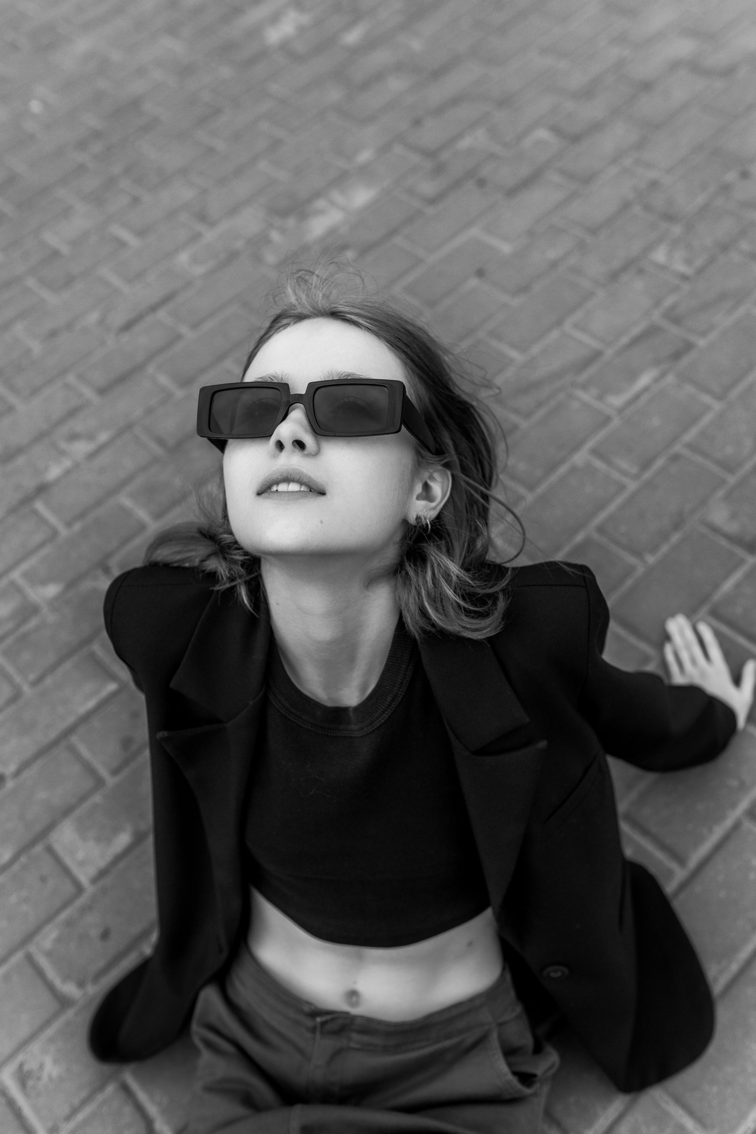 Девушк в черных солнцезащитных очках и блейзере сидит на кирпичном тротуаре, глядя вверх с безмятежным выражением лица, создавая непринужденную, стильную атмосферу.