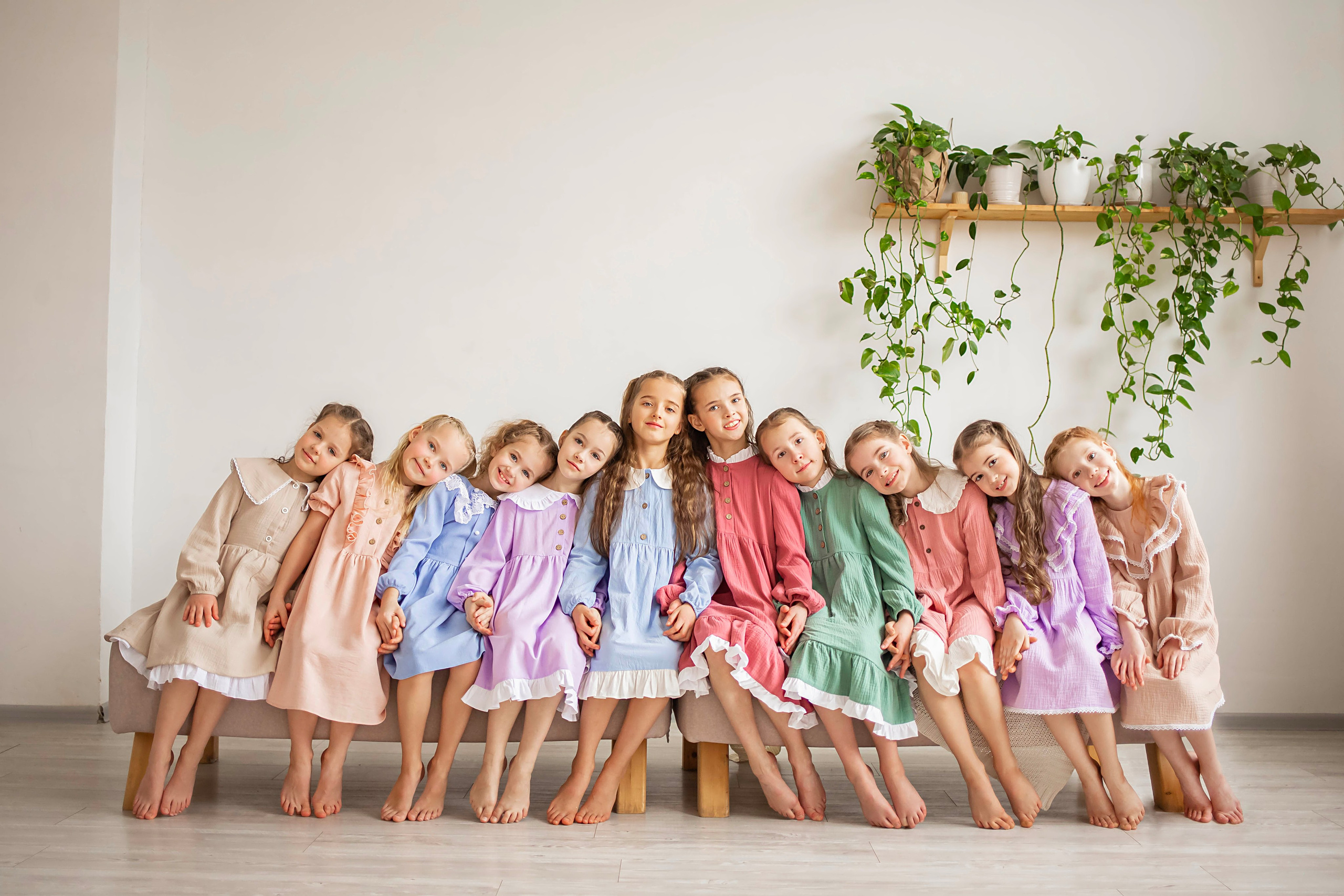 Детская съемка группы детей на разноцветных платьях в постельных тонах на фоне светлой стены и растений