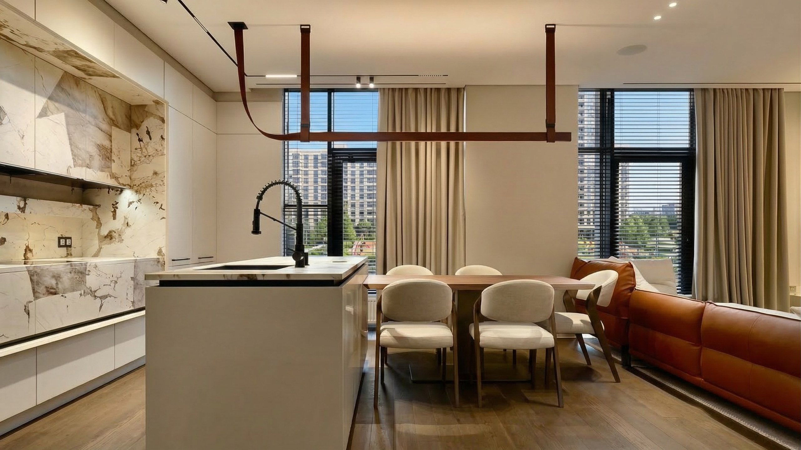 Современная кухня-гостиная с белым островом, мраморным фартуком, деревянным столом и уникальным линейным светильником на кожаных ремнях.