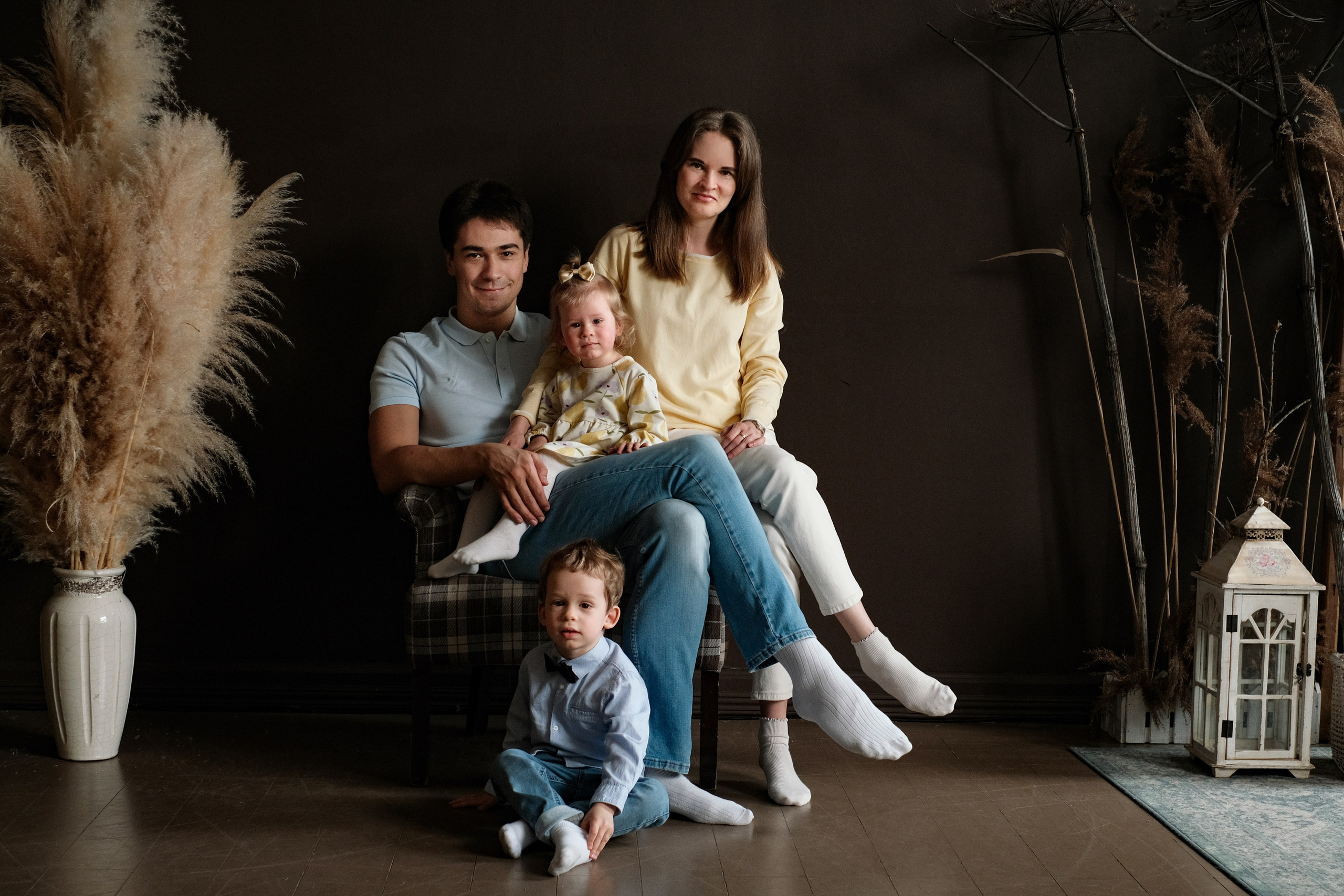 Семейная фотография в студии. Мама, папа, дочь сидит на коленях у папы, сын сидит в ногах у папы.
