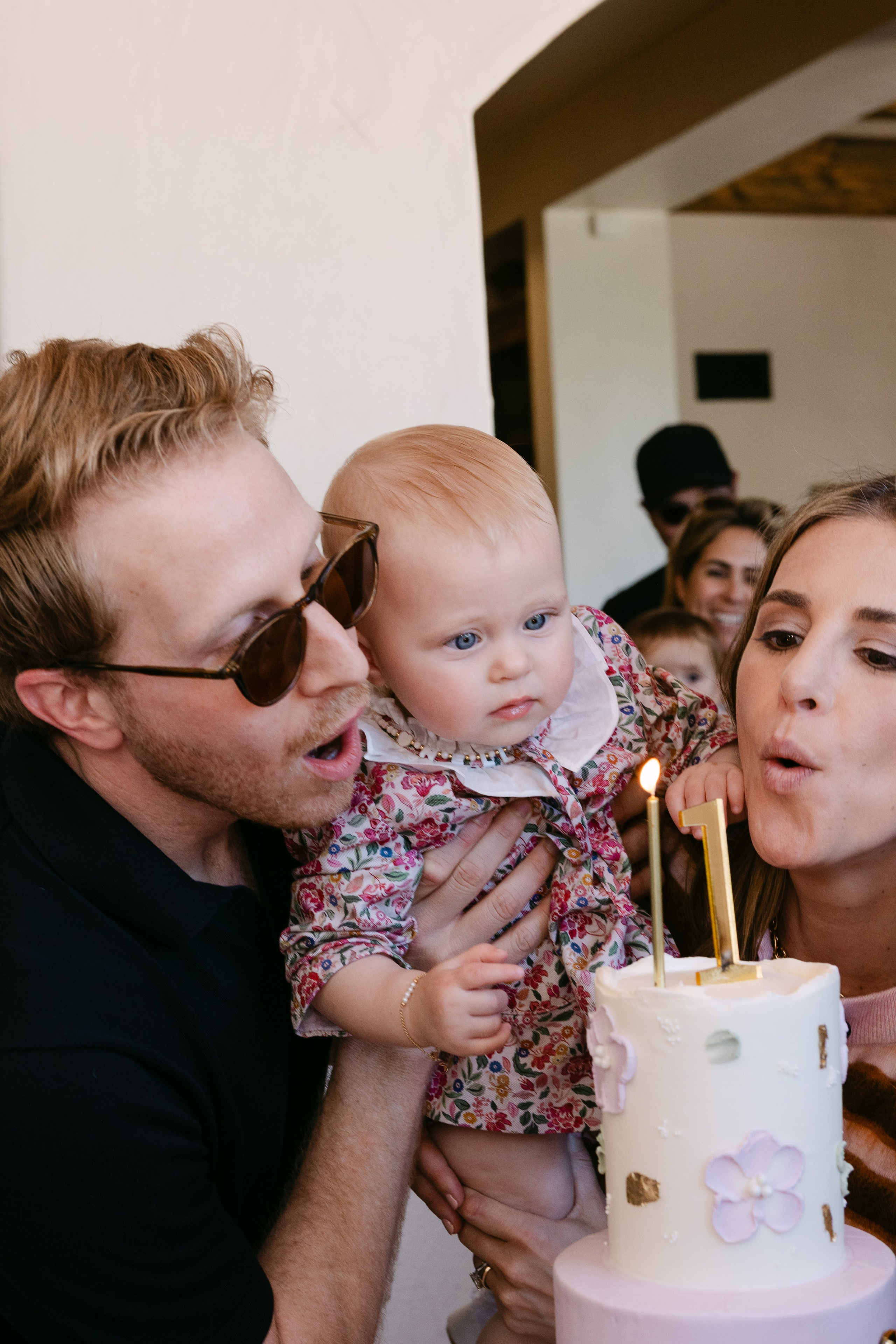 Mom and dad holding baby girl in a dress and helping her to blow candles on her big 1st birthday cake