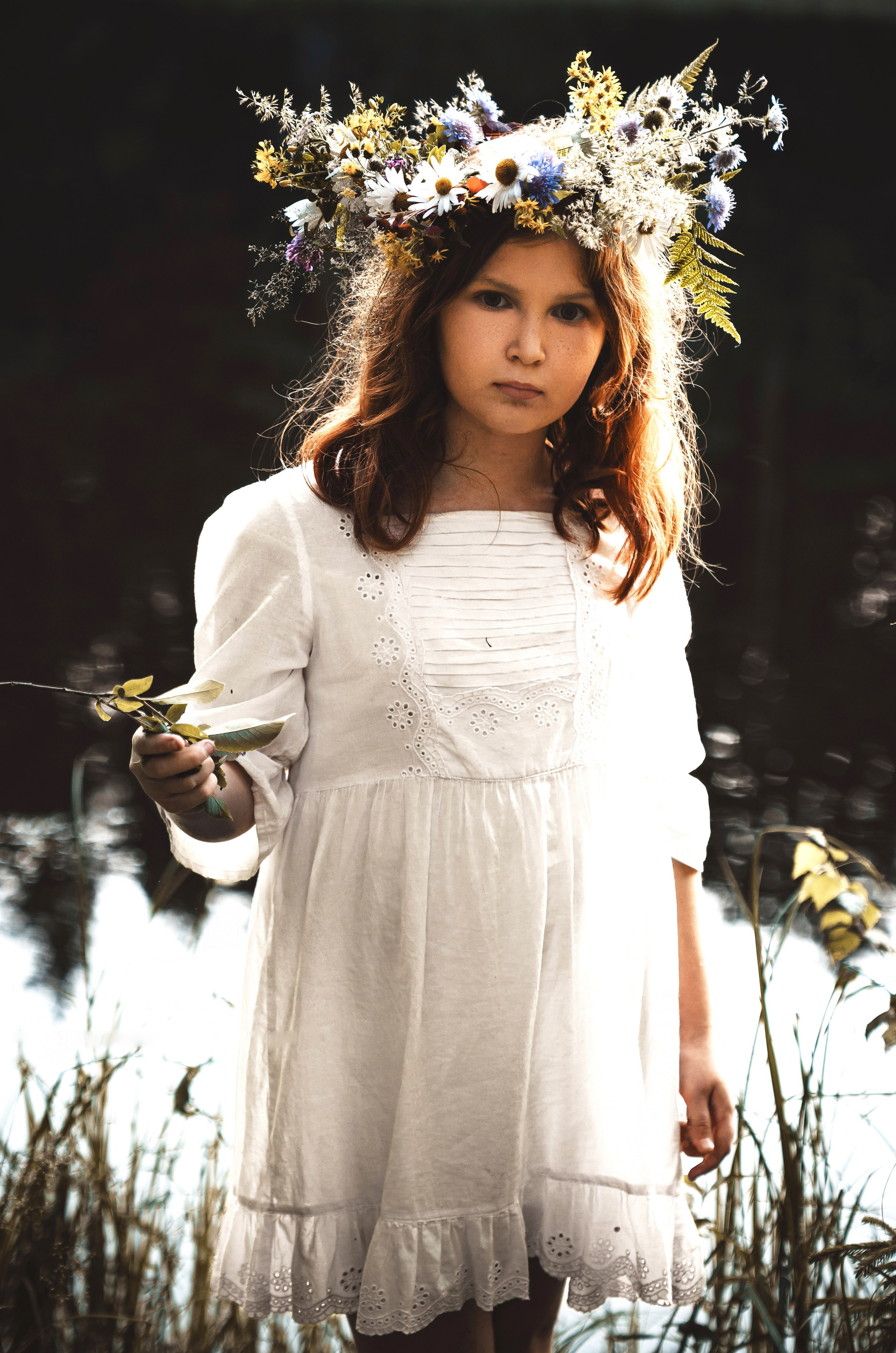 Девочка с рыжими волосами в венке из полевых цветов и папоротника держит веточку в лесу.