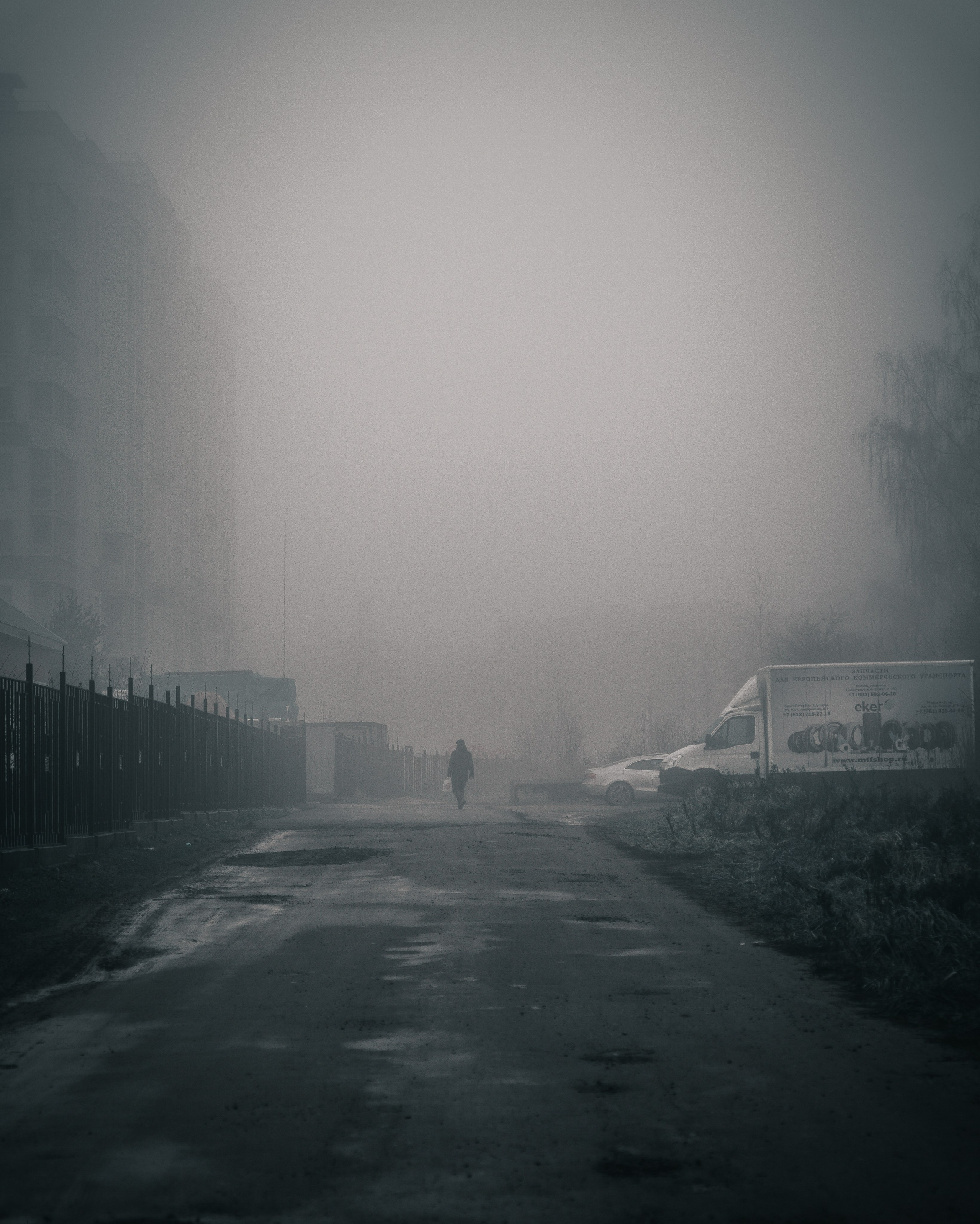 Одинокая фигура с пакетом идёт по заснеженному двору в густом тумане, панельные девятиэтажки Санкт-Петербурга, меланхоличная уличная сцена, фоторепортаж Евгения Мальцева.