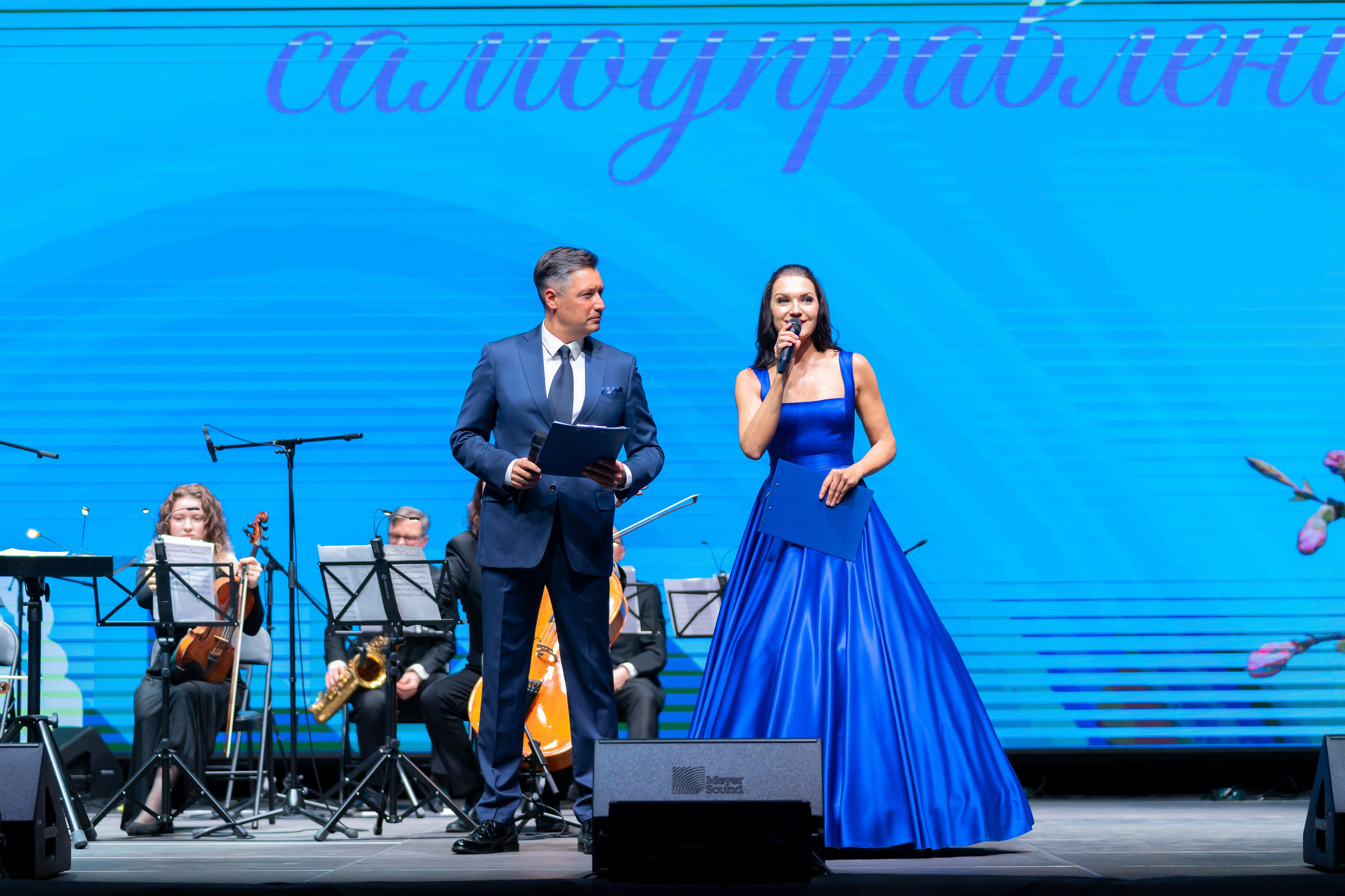 Ведущие концертной программы: мужчина в синем костюме и женщина в роскошном синем платье на сцене с оркестром. День местного самоуправления, Санкт-Петербург. Фото Евгения Мальцева.