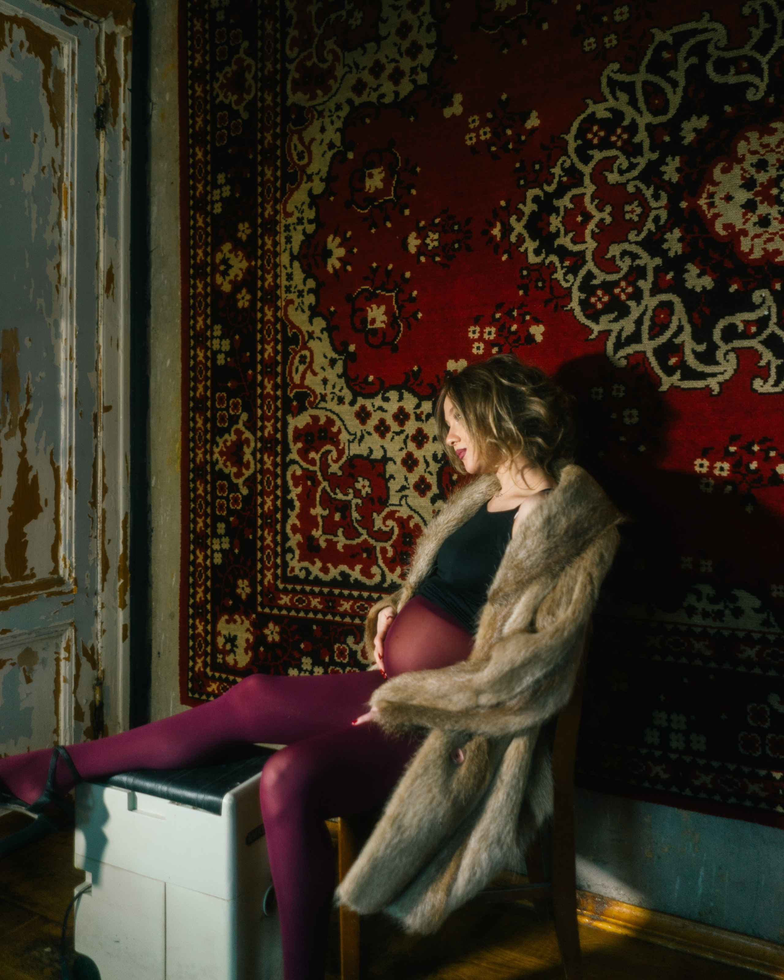 Беременная в шубе и бордовых (винных) колготках на фоне ковра на стене — дерзкая ретро-фотосессия беременности в фотостудии СПб в стиле девяностых.