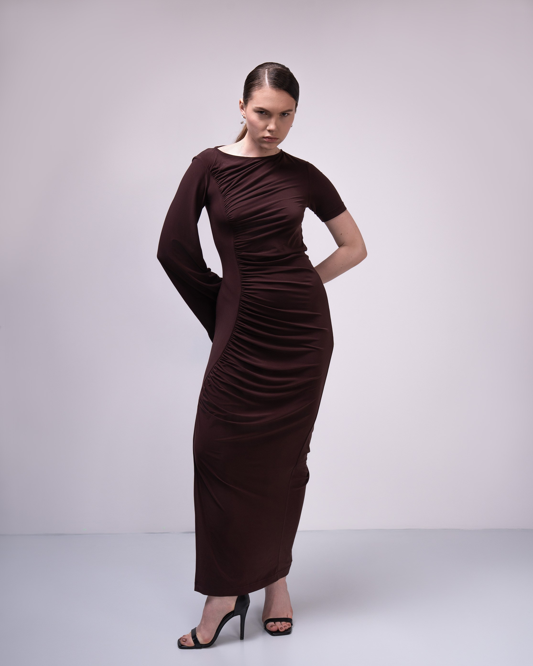 Фото товара для каталога  Ozon или Wildberries.  На фотографии изображена модель, демонстрирующая вечернее платье. Платье выполнено в темно-коричневом цвете и имеет облегающий силуэт. Оно украшено драпировкой на груди и рукавах, что придает ему элегантный и изысканный вид. Модель стоит на нейтральном фоне, что позволяет сосредоточиться на изделии. На ногах у модели черные туфли на высоком каблуке, что подчеркивает длину платья и делает образ завершенным.
