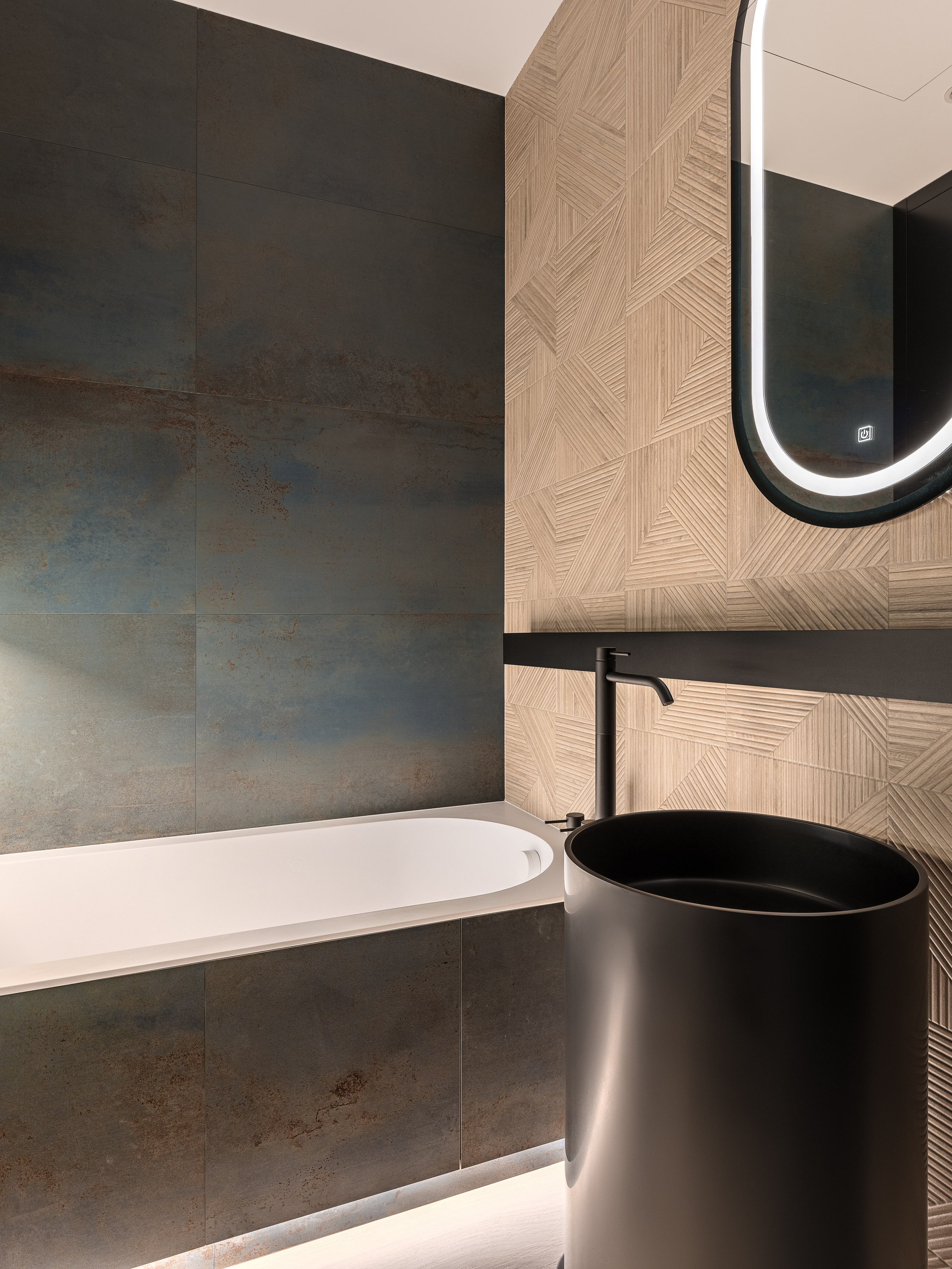 Современная ванная комната с акцентной стеной из плитки под металл, геометрической бежевой отделкой, подвесным зеркалом с подсветкой, черной цилиндрической раковиной и минималистичными смесителями. Дизайнеры Ренат Байчуков и Алеся Кириенко, стилист Екатерина Ракитина, фотограф Сергей Денисенко.