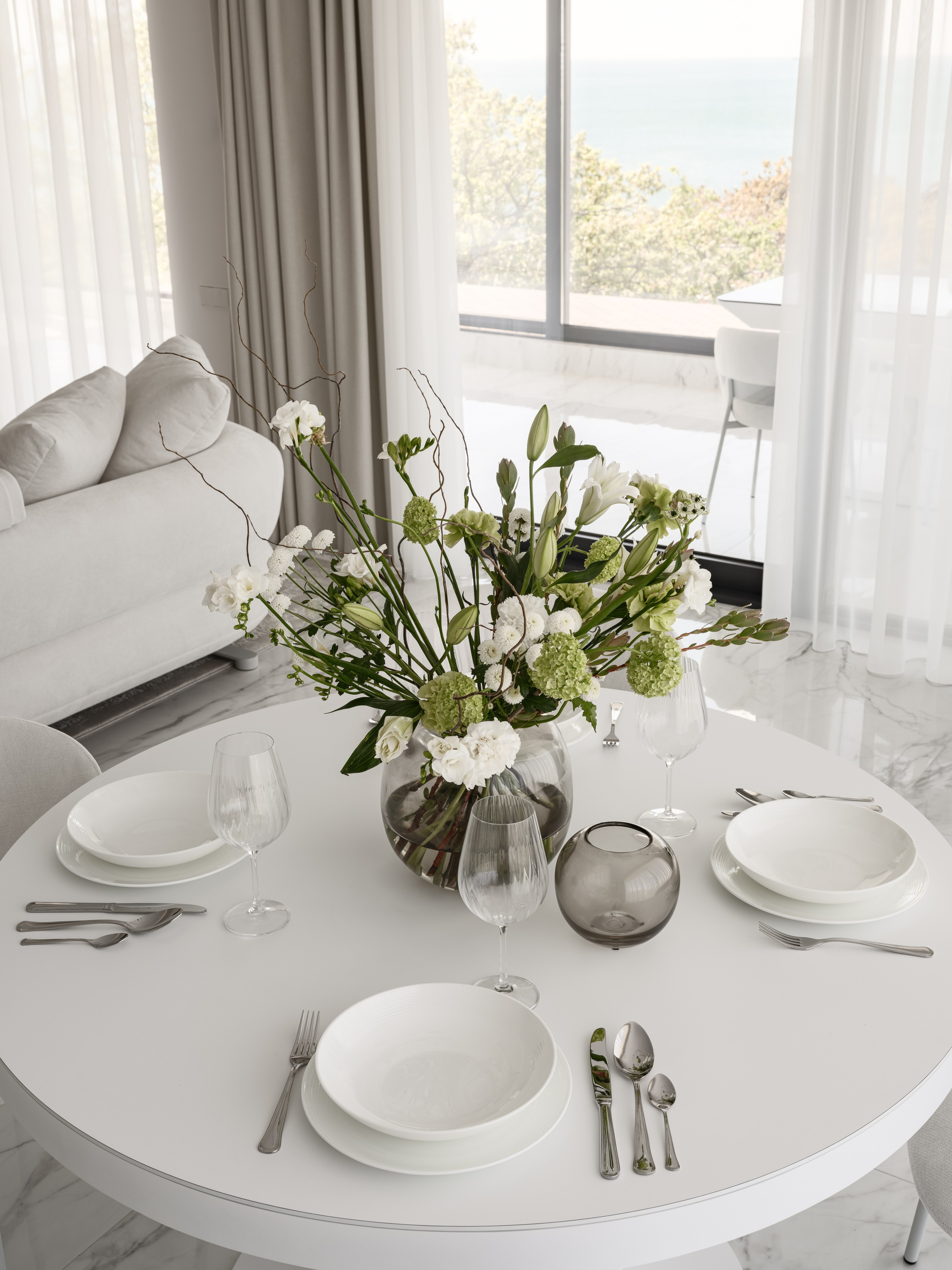 Круглый обеденный стол в светлой гостиной с сервировкой на четыре персоны и букетом белых цветов в стеклянной вазе, панорамные окна с видом на море и мягкий диван — интерьер от дизайнера Алёны Боярской, стилист Наталья Якушева, фото Сергей Денисенко.
