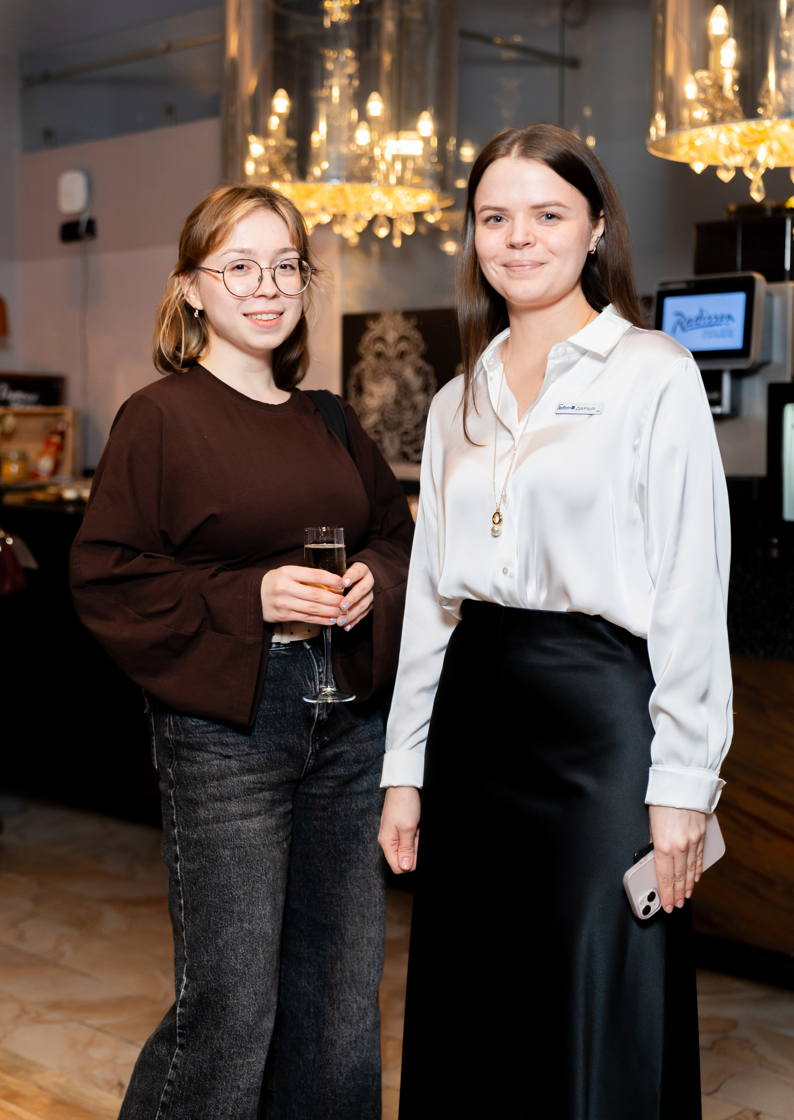 Две девушки на мероприятии: в коричневом свитере с шампанским и сотрудница отеля в белой блузке. Фото Евгения Мальцева, Radisson Sonya.