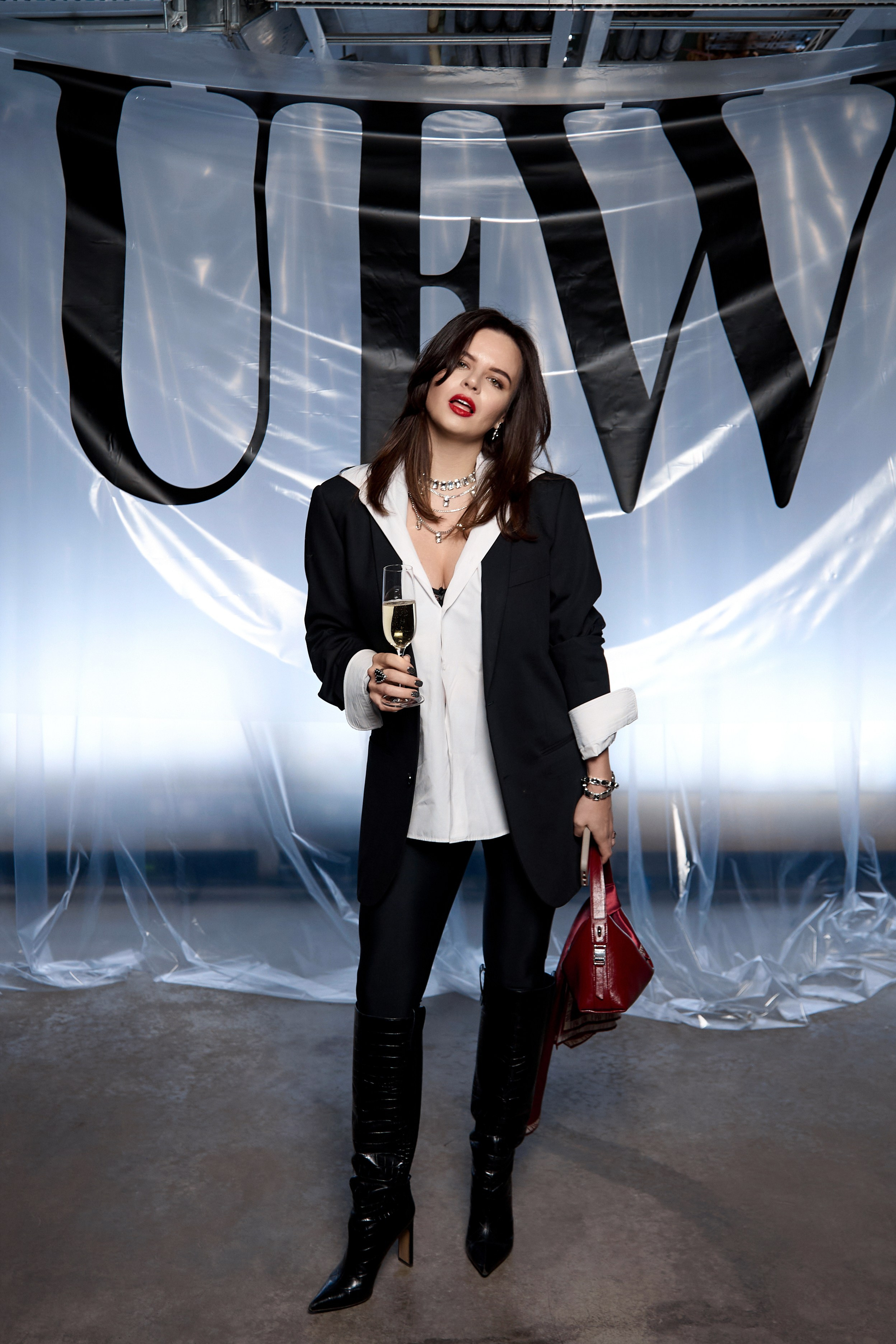 Улыбающаяся гостья в белом вечернем платье и с бокалом шампанского на фоне модного показа.