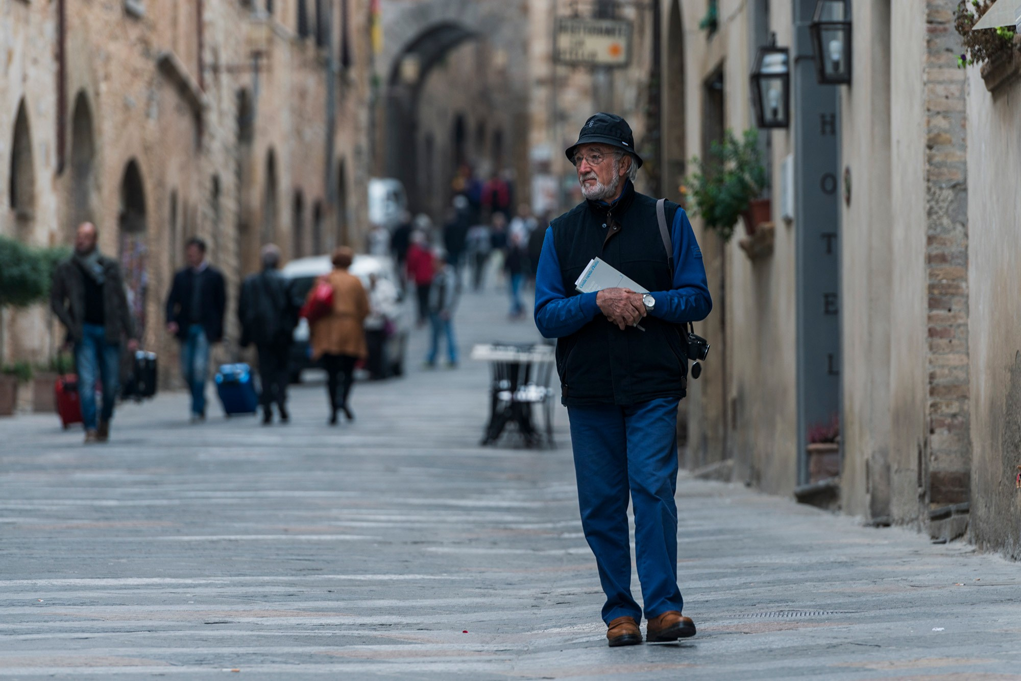 Пожилой мужчина в черной шляпе и синем жилете идет по узкой каменной улице в историческом центре Пьенцы, Италия, во время сессии уличной фотографии.