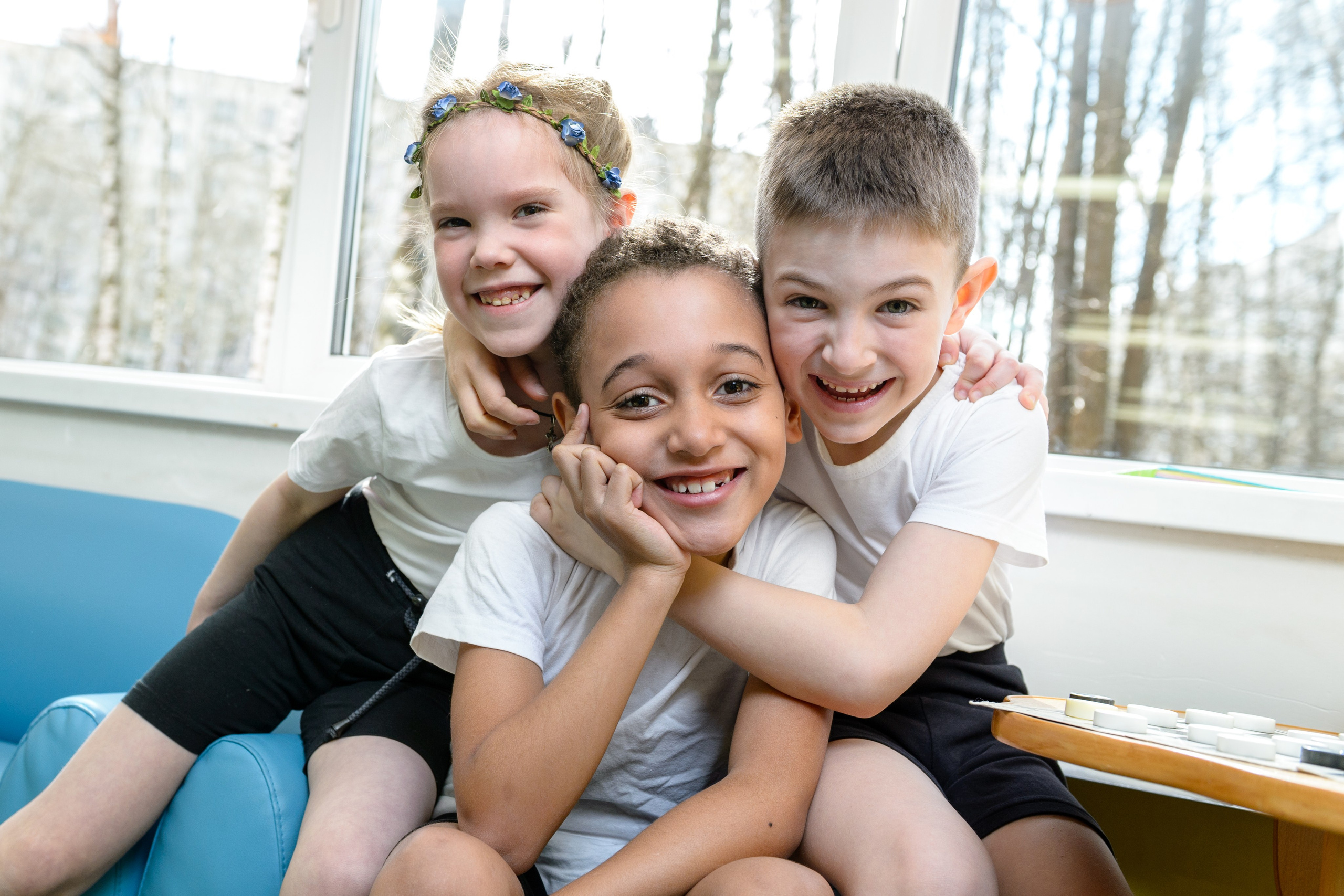 Трое детей в спортивной форме обнимаются у окна и смеются в дружеском кадре в помещении.